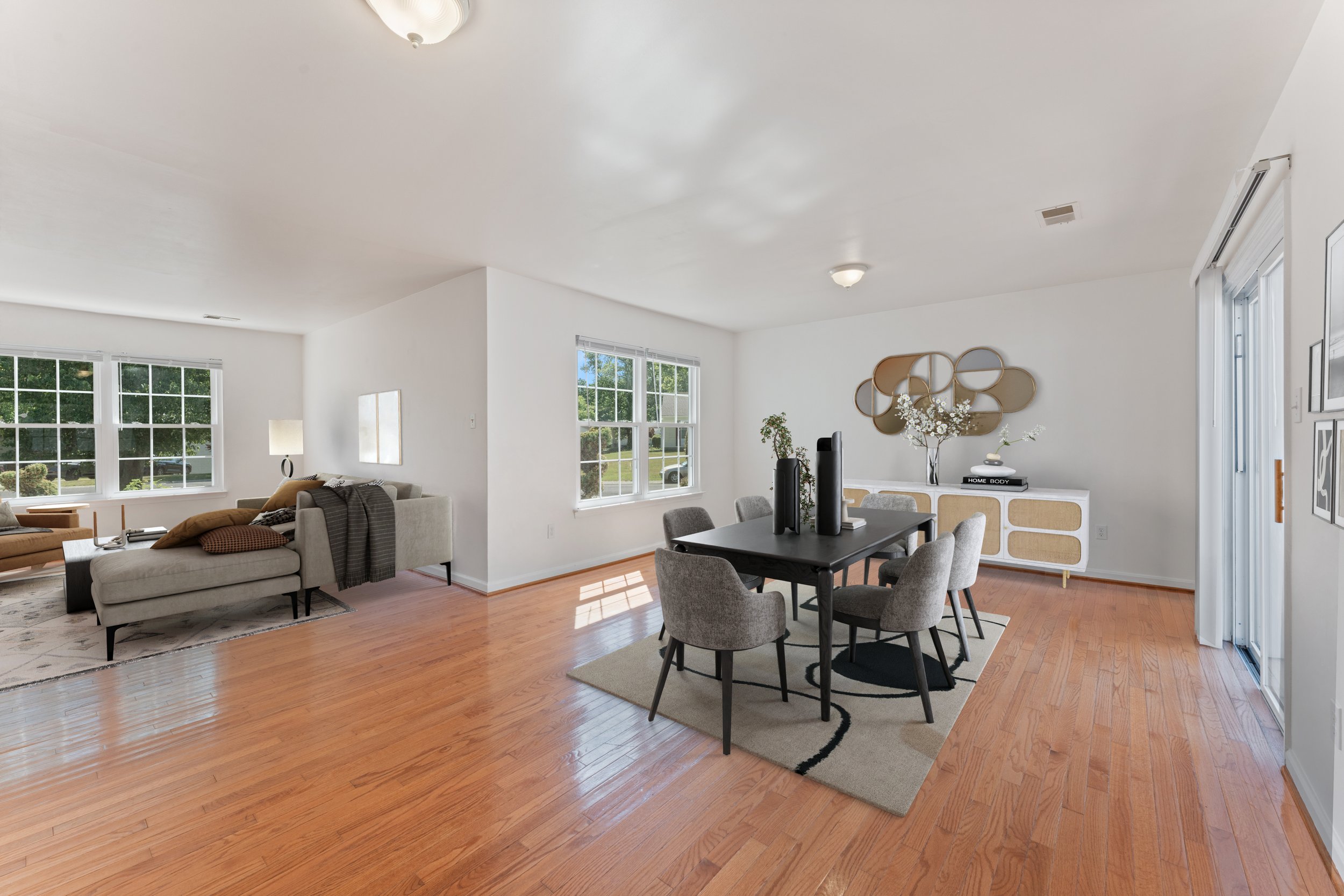 Open living and dining room with white walls, hardwood floors, windows, and contemporary furniture, including a black dining table with six chairs, a white sideboard, decorative wall art, and a lounge area with a sofa, chairs, and a rug.