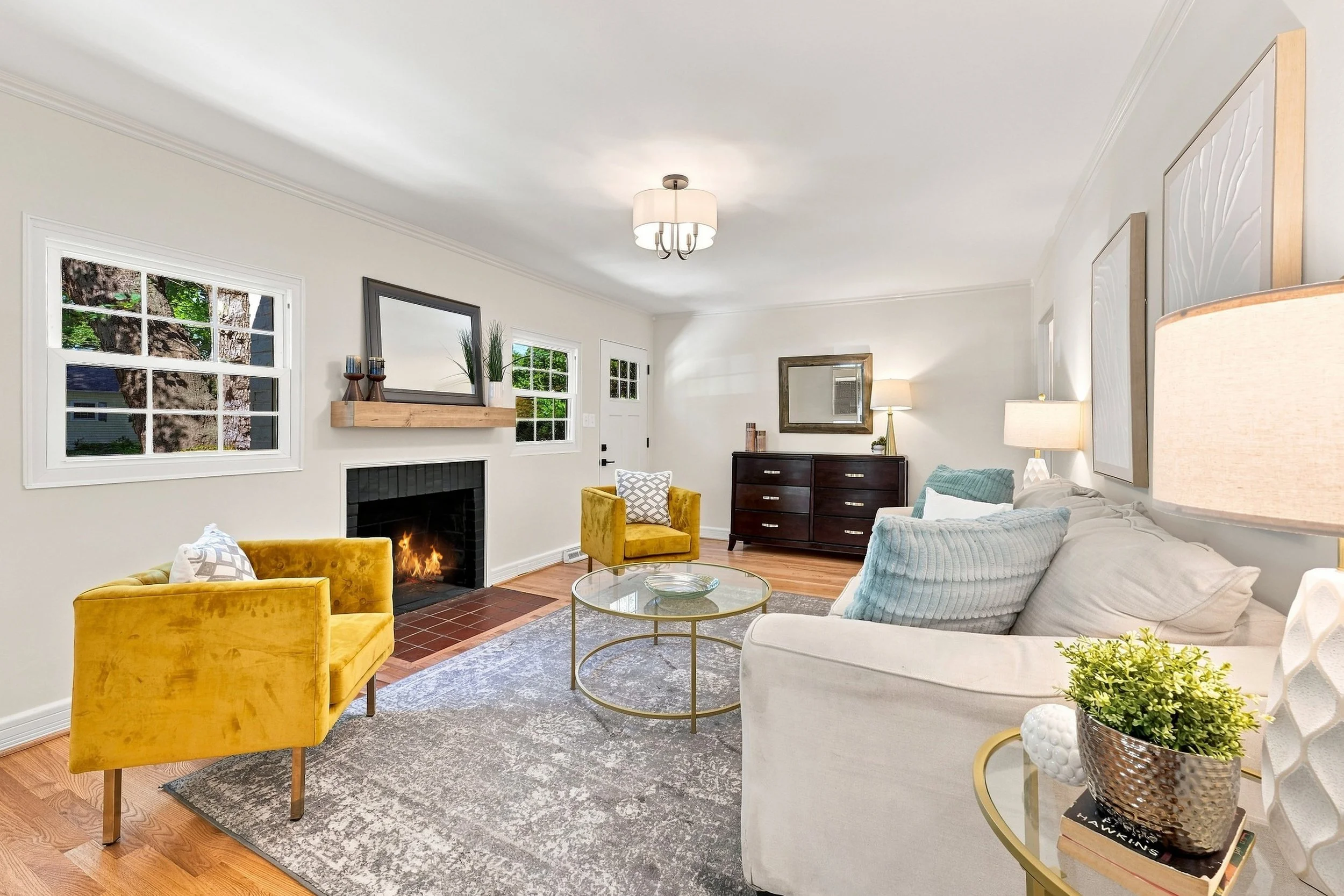 Staged living room with a lit fireplace, mustard velvet chairs, and daylight through white-trimmed windows
