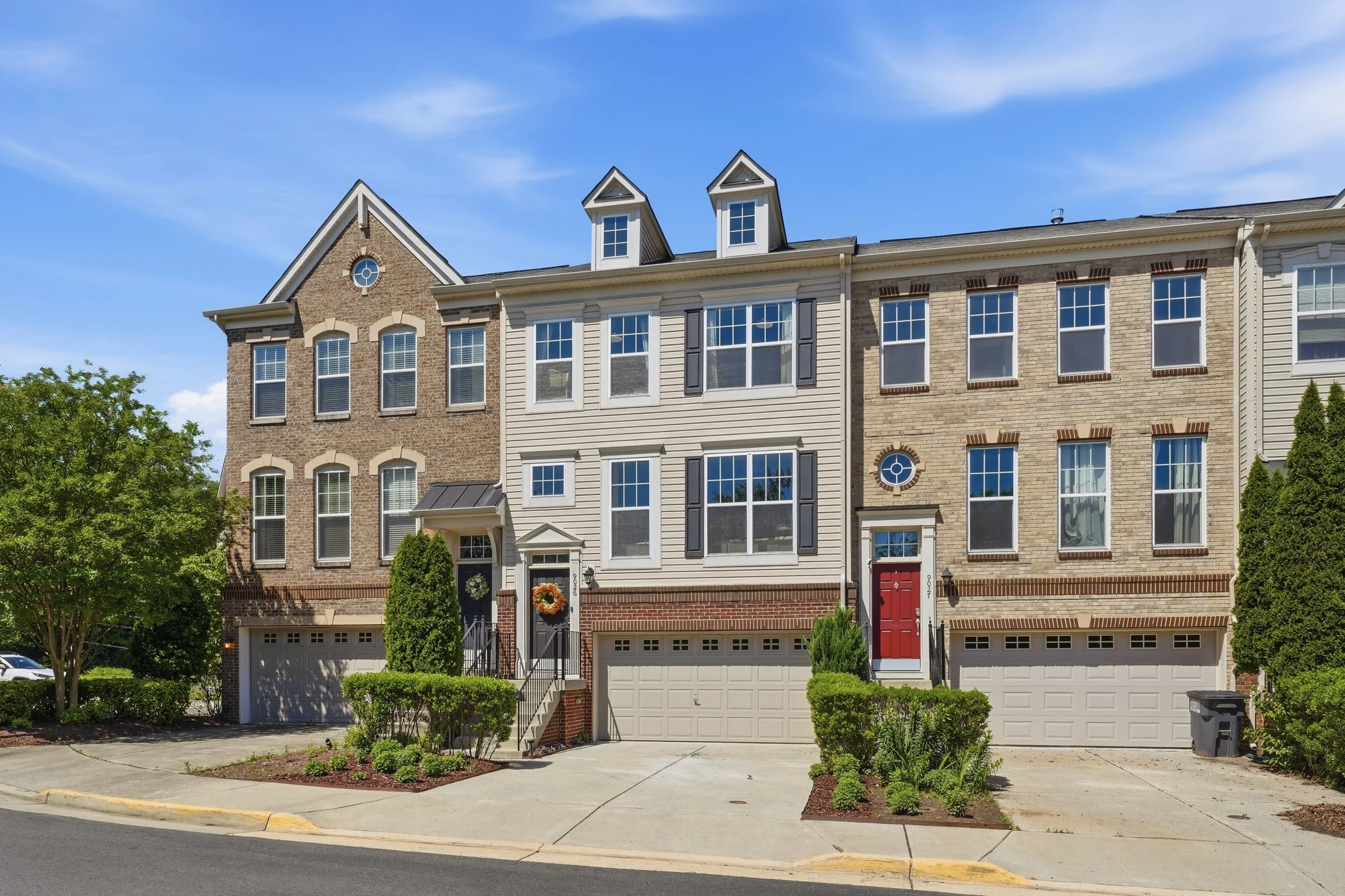 Three attached townhomes, brick end units flanking a tan-sided center unit with two dormers, each with a two-car garage, under a blue sky with light clouds.
