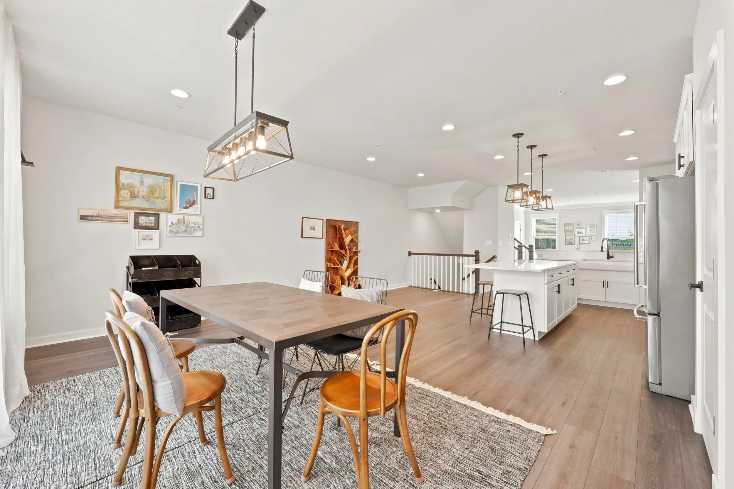 Dining area with a wood table and bentwood chairs, looking back toward a white kitchen island, stainless refrigerator and stair railing on an open main level.