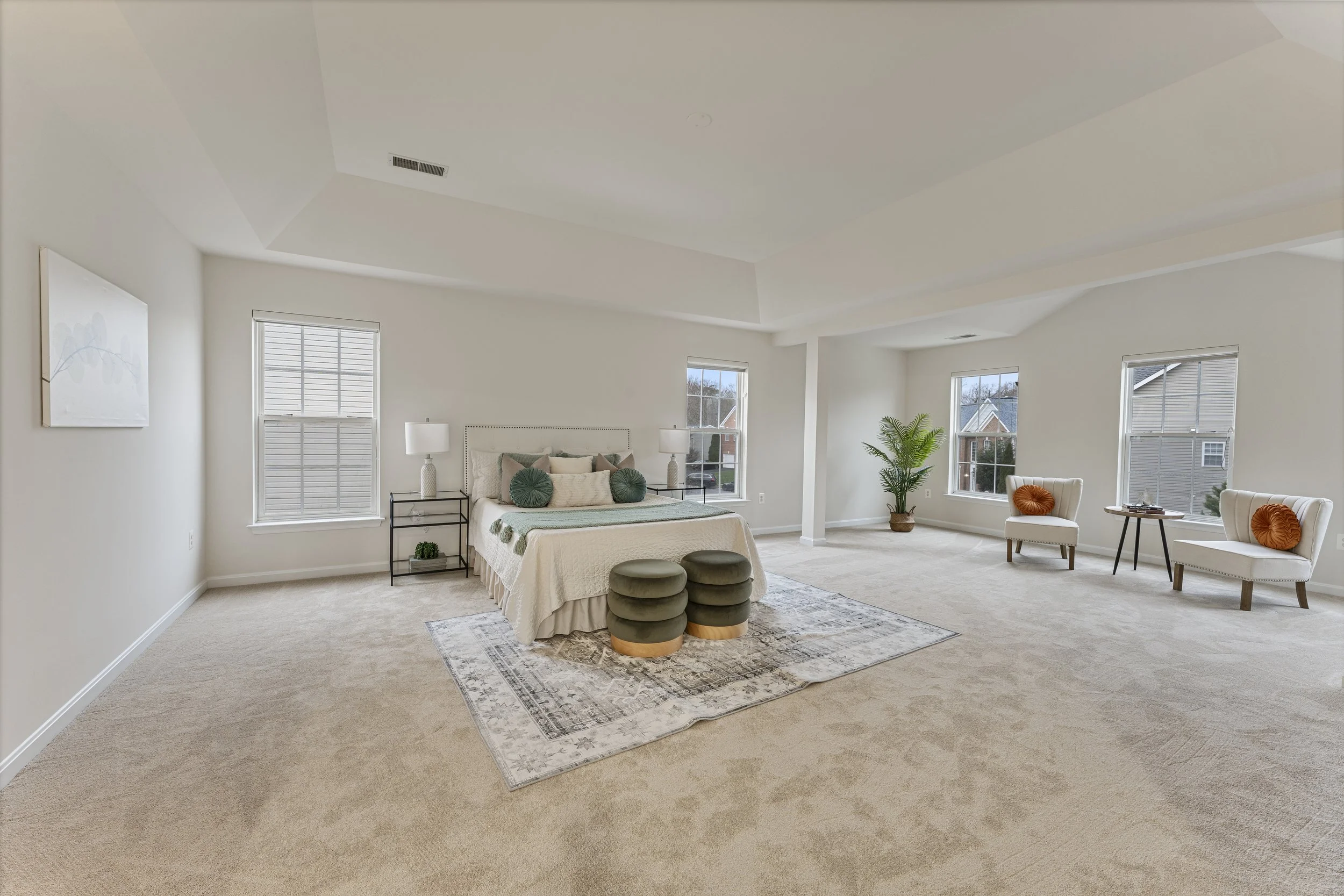 Spacious bedroom with beige carpet, white walls, and several windows, featuring a bed with pillows, side tables, a large potted plant, and seating area with armchairs and side tables.