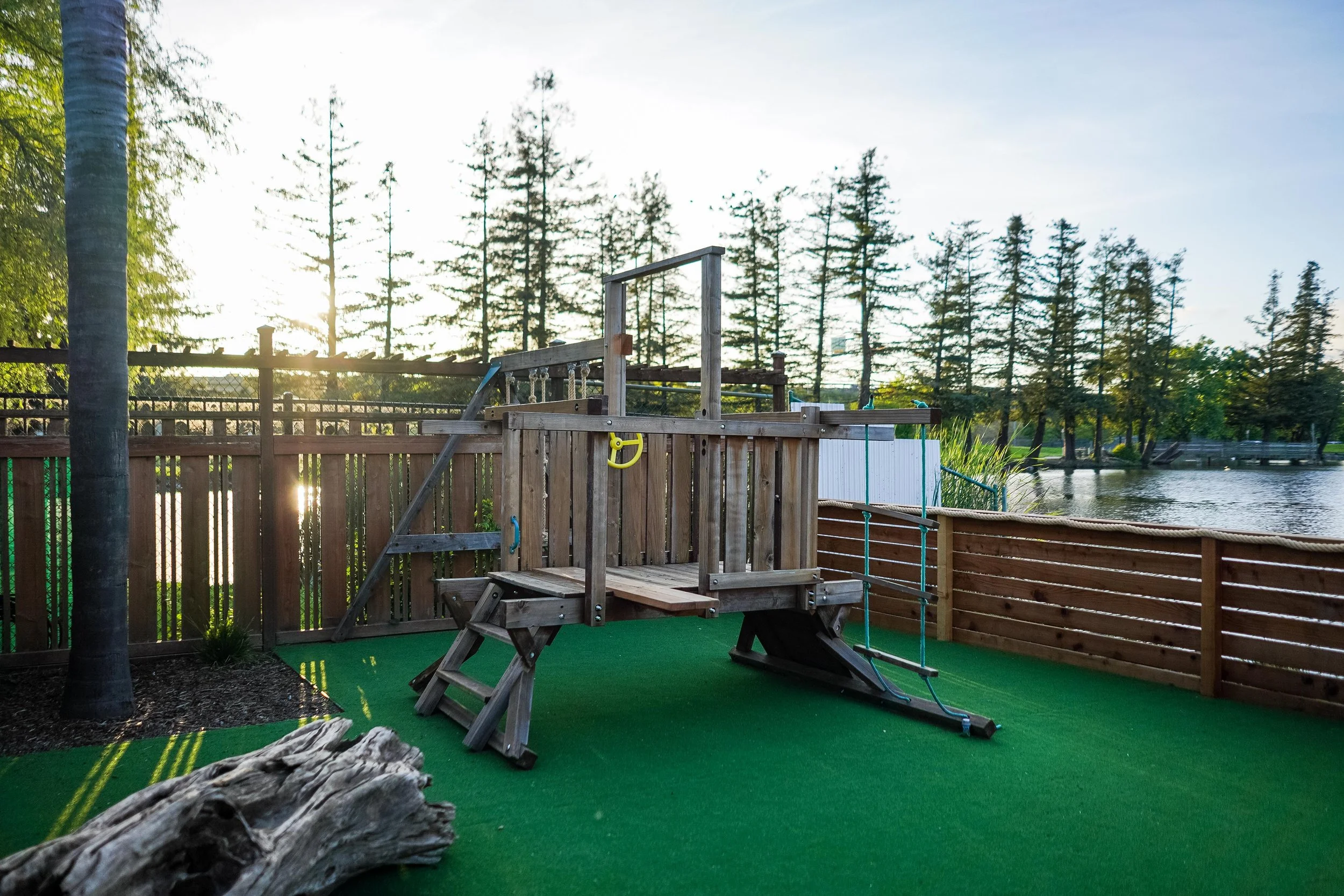 Backyard scene with a wooden play structure on a green artificial turf, a lake with trees in the background, and a fence.