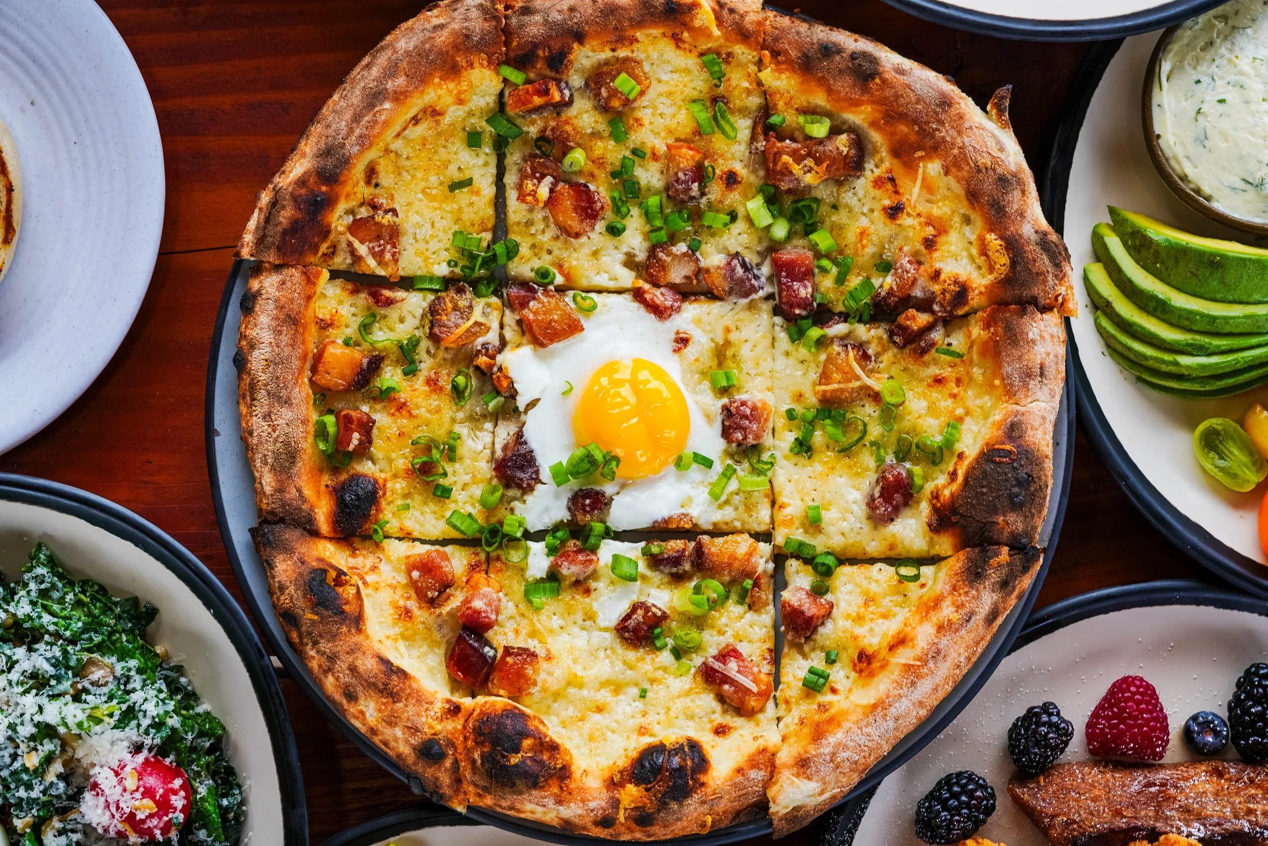 A pizza with a fried egg, chopped green onions, cheese, and bacon pieces on a black plate. There are sides of avocado slices, a bowl of creamy dip, a plate of fruit, and a leafy green dish around it, all on a wooden table.