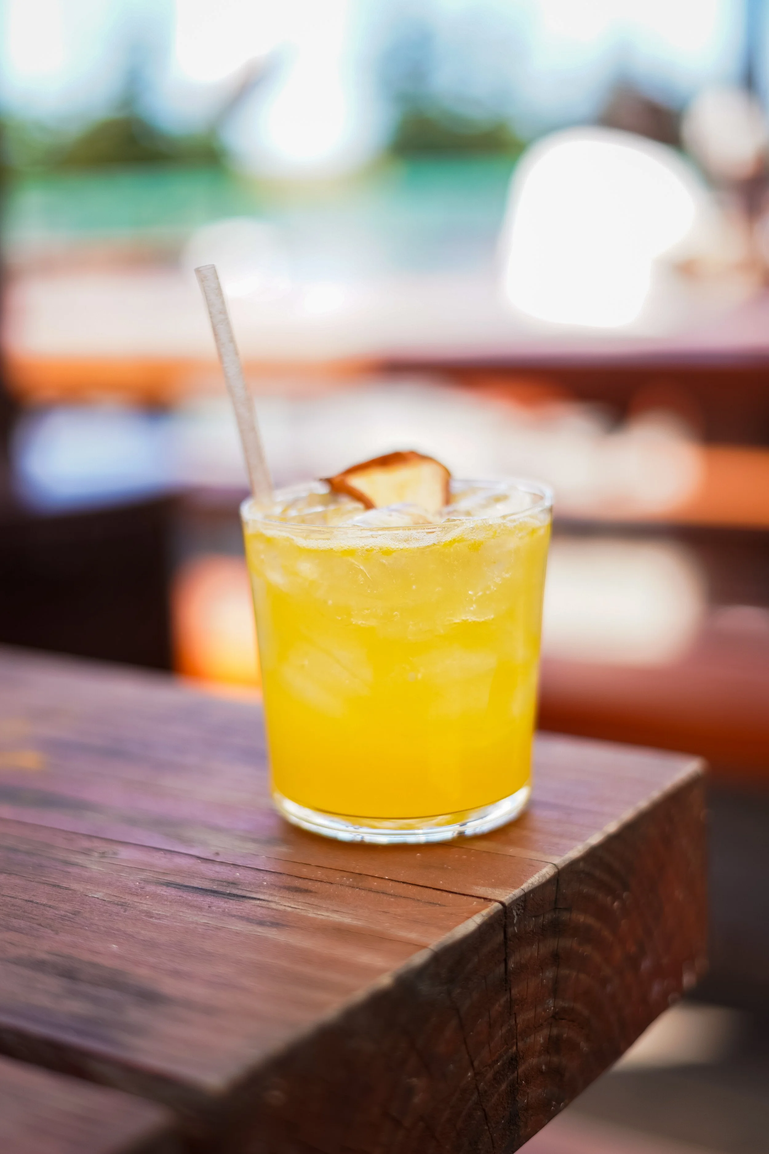 A glass of yellow citrus drink with ice and a lime wedge, placed on a wooden table at a sunny indoor setting.