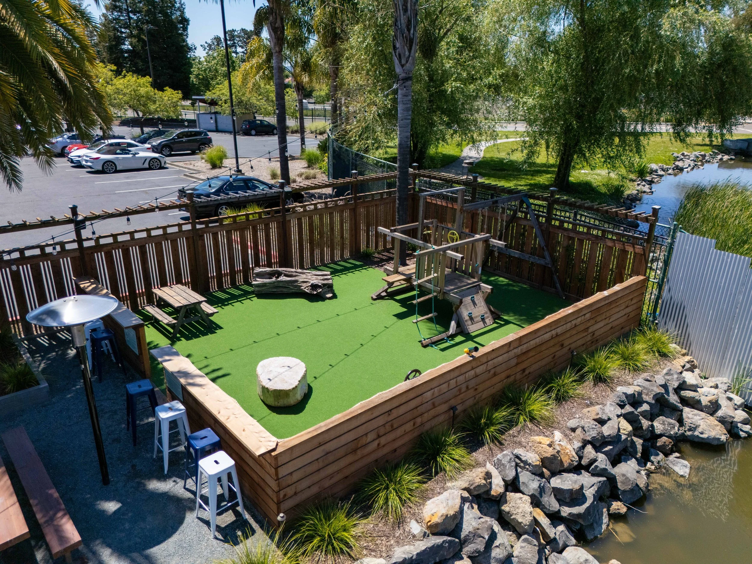 A backyard with a fenced patio area and a small pond with rocks and grass. There are trees and parking lot in the background.