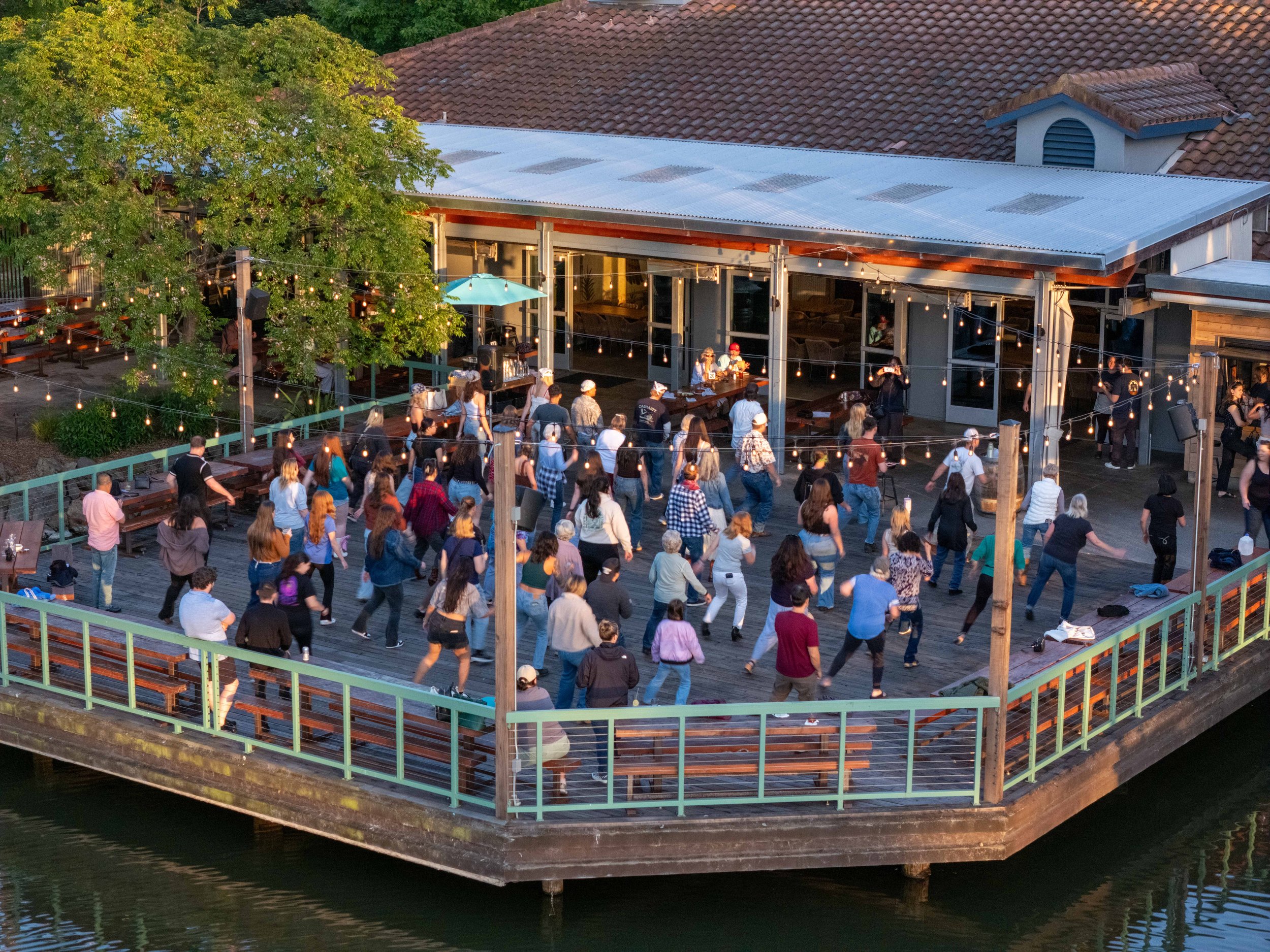 An outdoor event on a wooden deck with many people dancing and socializing. There are string lights hanging and a few tables with umbrellas, surrounded by green trees and a building with a tiled roof.