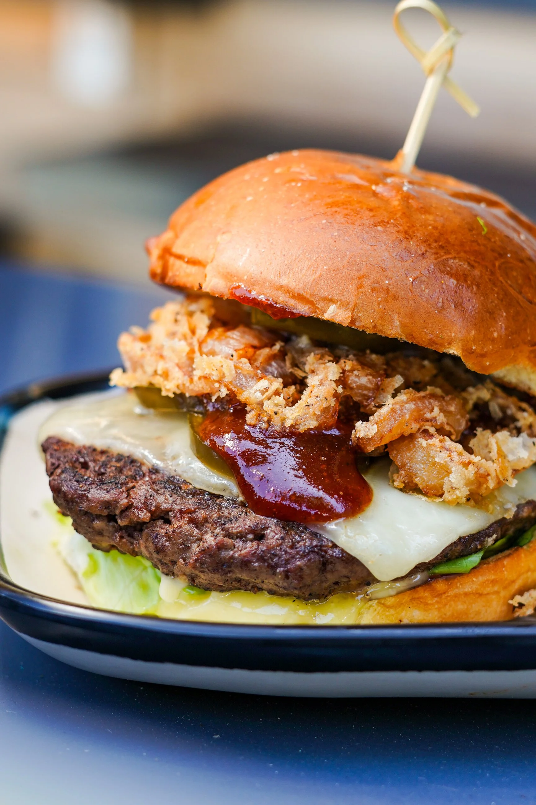 Close-up image of a cheeseburger with a beef patty, melted cheese, fried onions, barbecue sauce, pickles, lettuce, and a toasted bun, served on a black and white bowl.