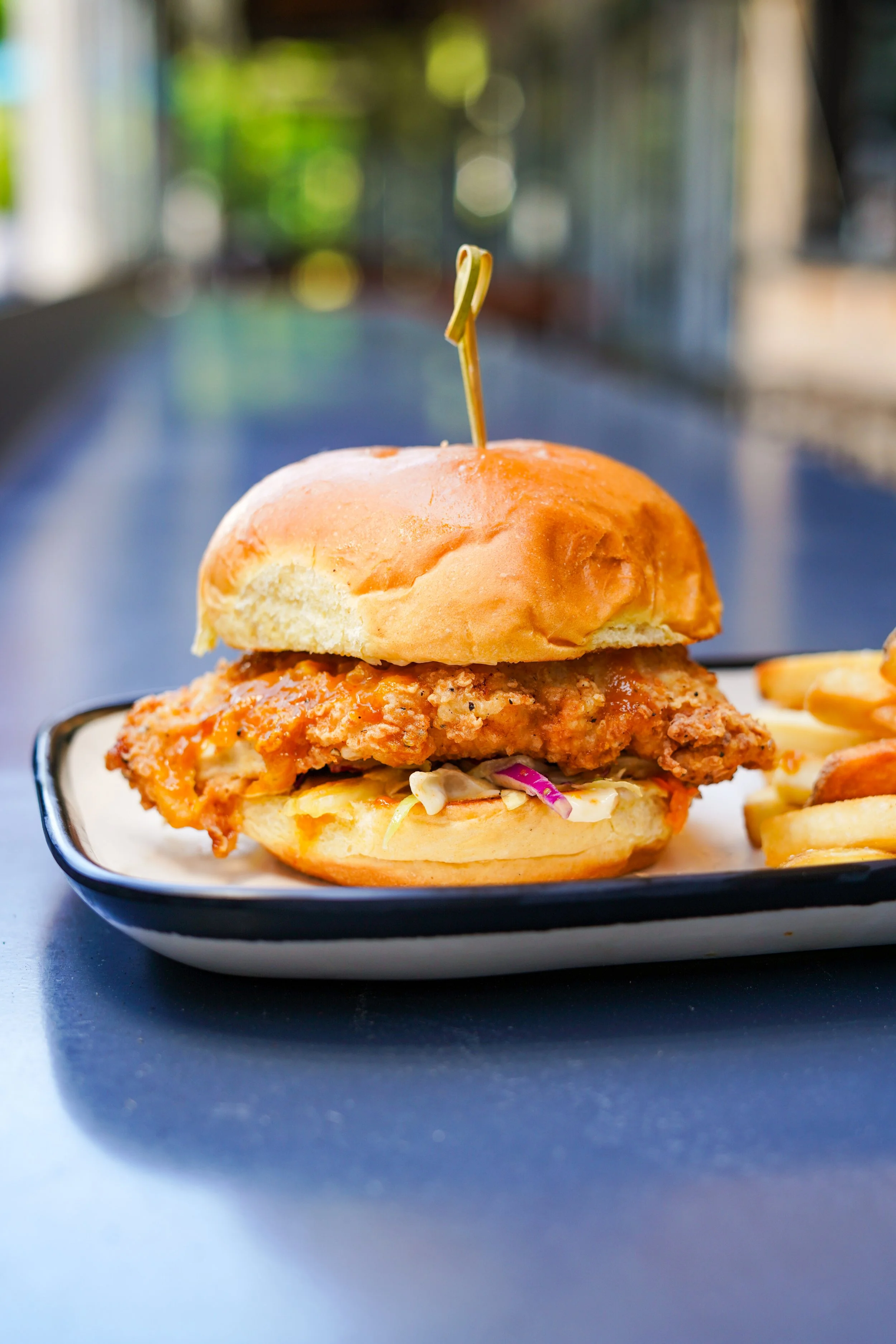Close-up of a fried chicken sandwich with coleslaw and pickles on a bun, served on a black and white rectangular plate, with a side of potato chips, on a dark table.