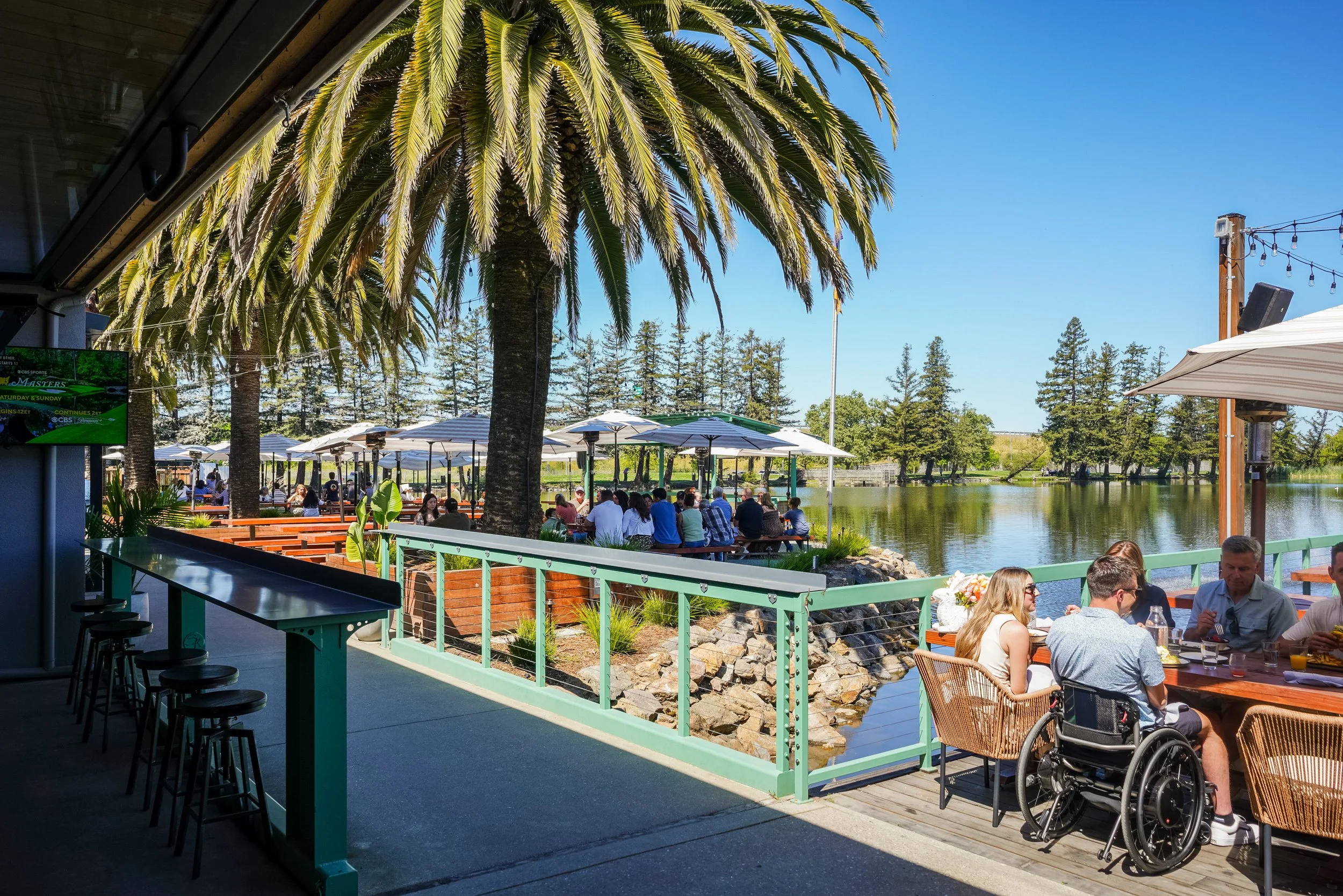 An outdoor dining area by a body of water with palm trees and people sitting at tables under umbrellas, sunny sky.
