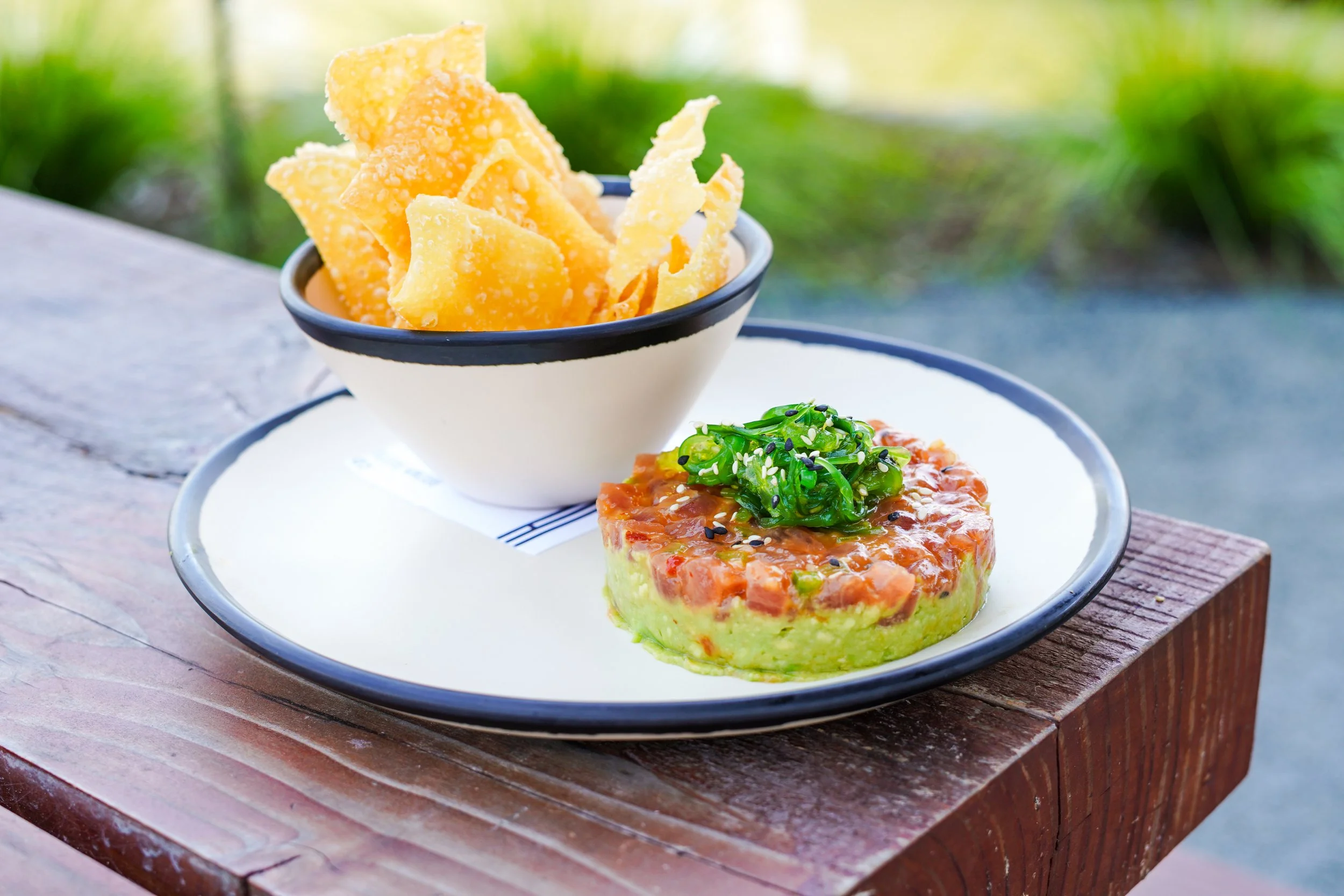 A bowl of crispy cheese chips on a plate, served alongside a piece of salmon topped with chopped green onions and black sesame seeds, on a wooden table outdoors.