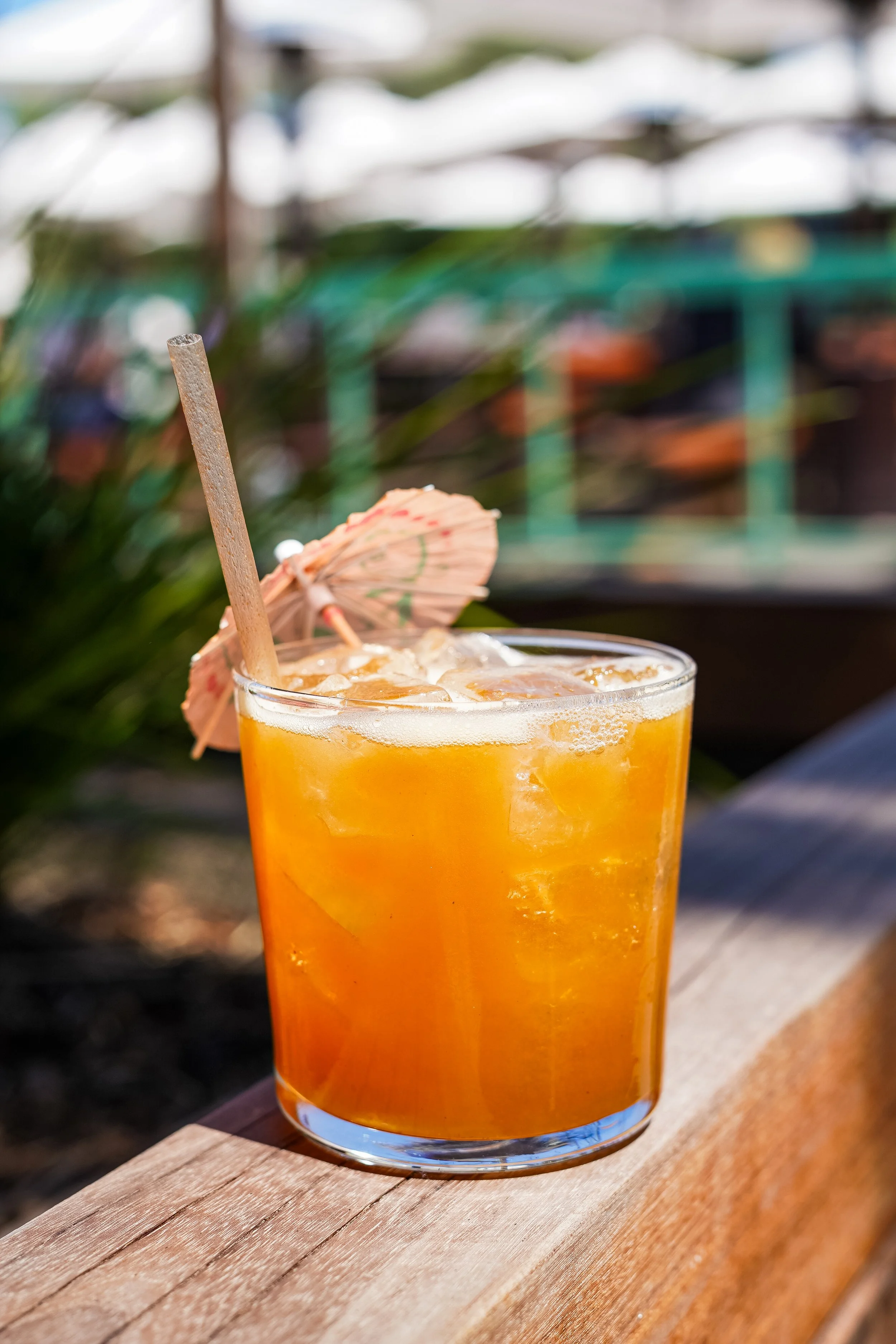 A glass of orange-colored beverage with ice, a paper umbrella, and a straw, placed on a wooden surface outdoors.