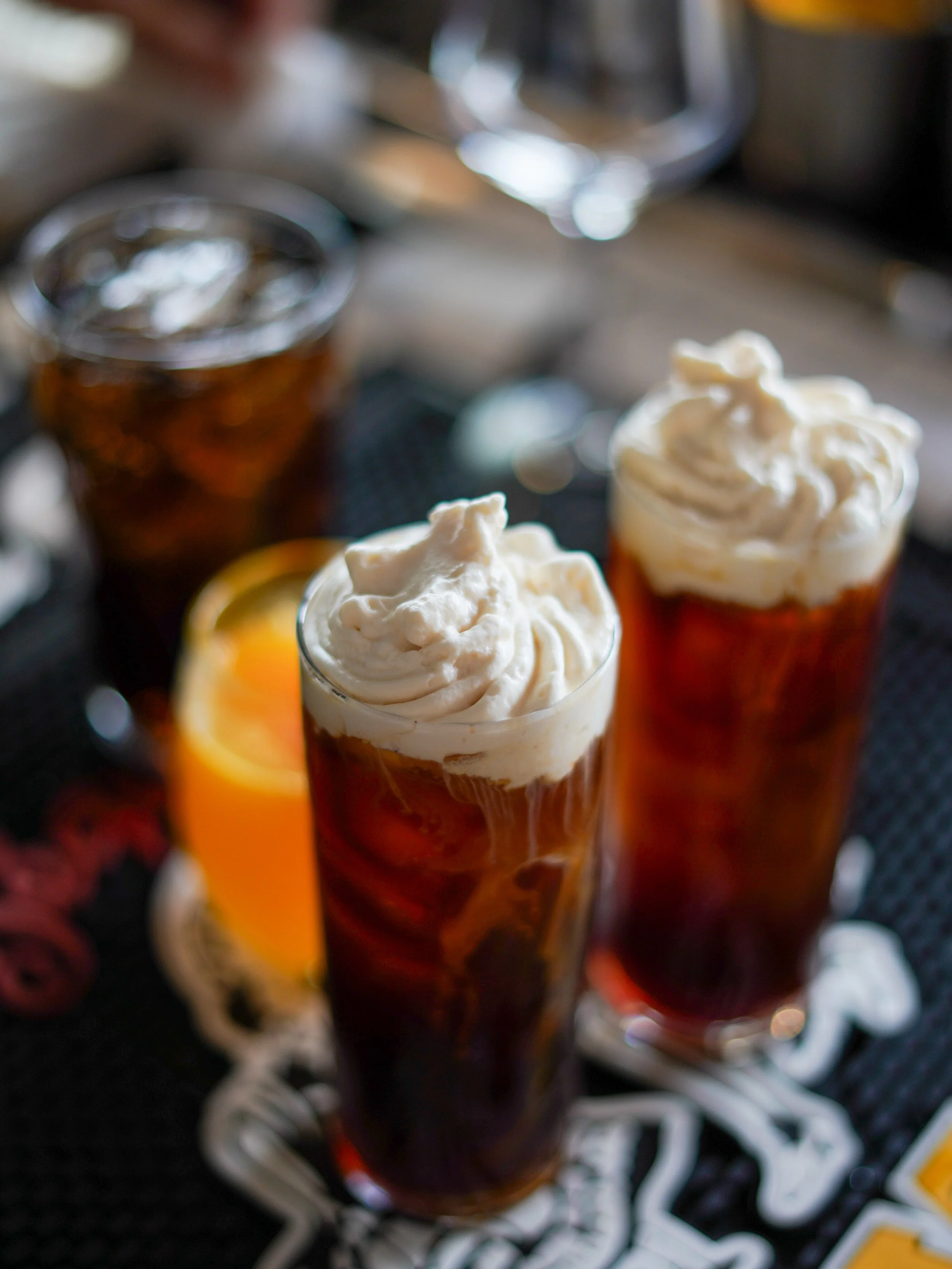 Three glasses filled with a dark beverage topped with whipped cream, placed on a decorative black and white table with a lemon slice in the background.