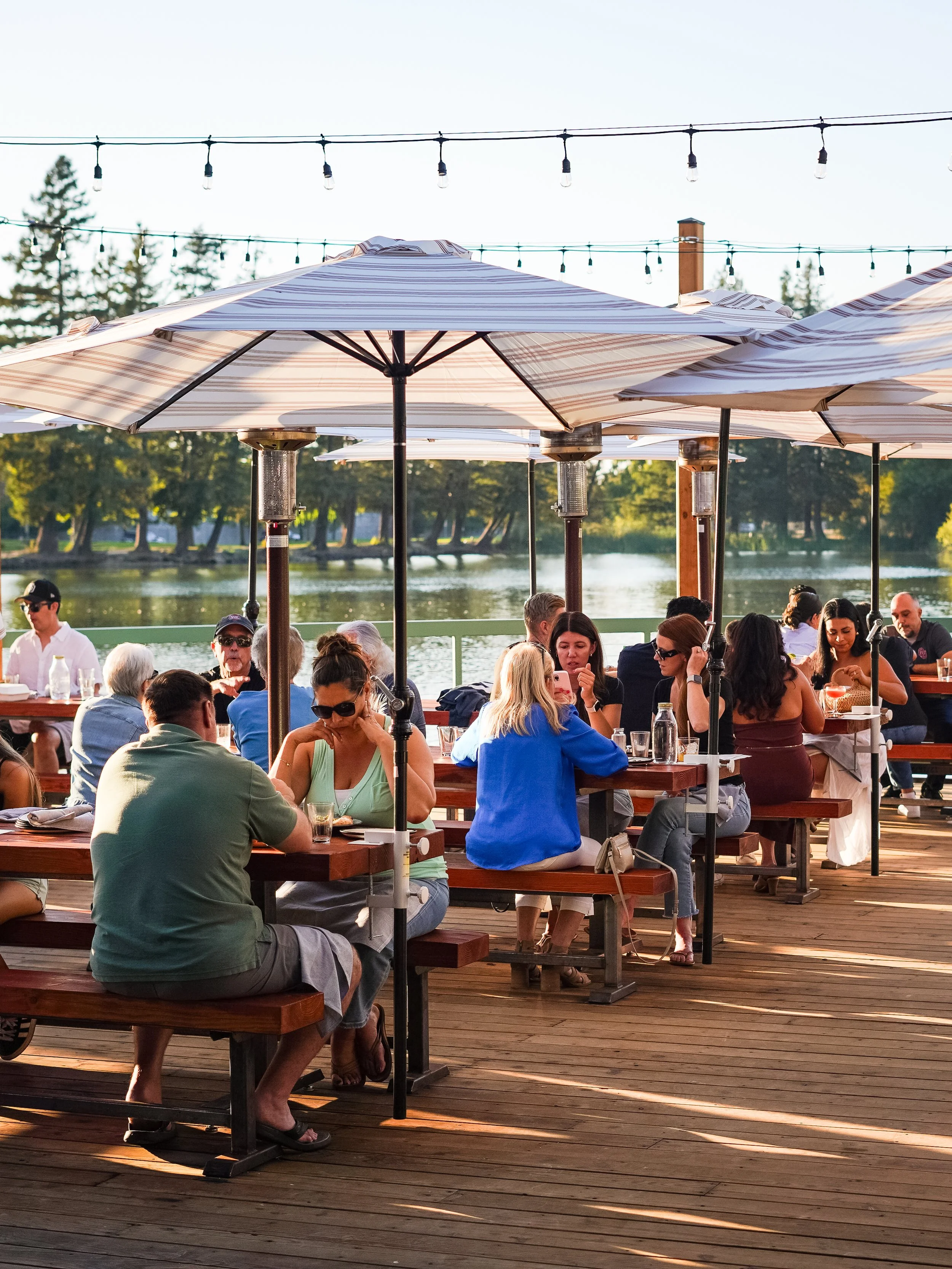 People dining under umbrellas on a wooden deck by a body of water with trees in the background.
