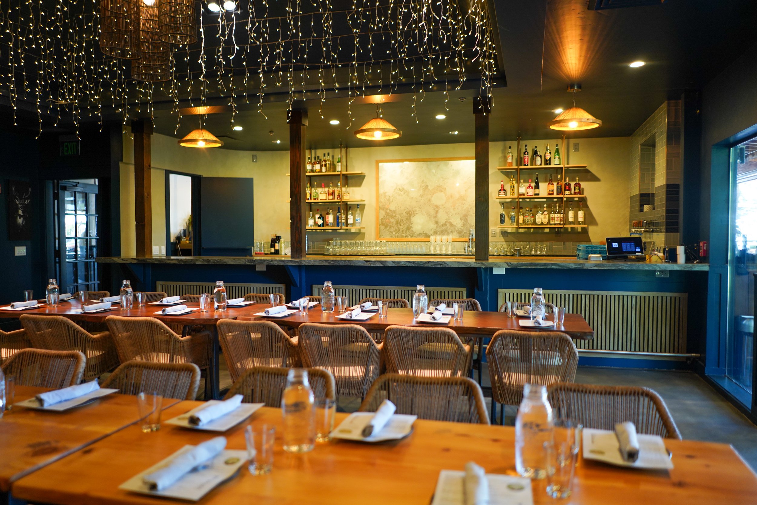 Restaurant with a long wooden table set with glasses, napkins, and small plates, surrounded by wicker chairs. In the background, a bar area with bottles of alcohol on shelves, warm pendant lighting, and decorative string lights hanging from the ceili
