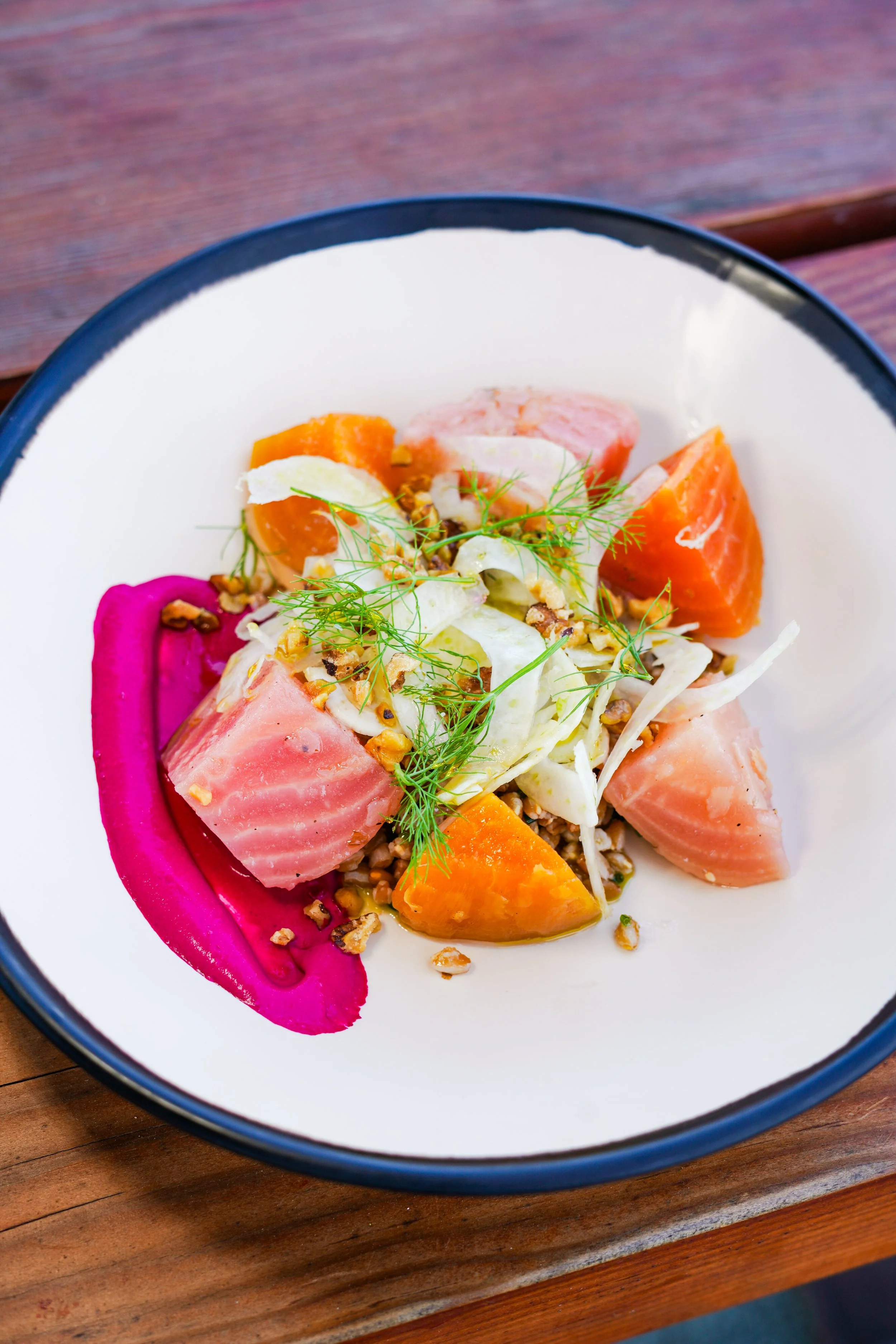 A bowl of colorful salad with pieces of salmon, butternut squash, pickled beet, and garnished with dill and chopped nuts.