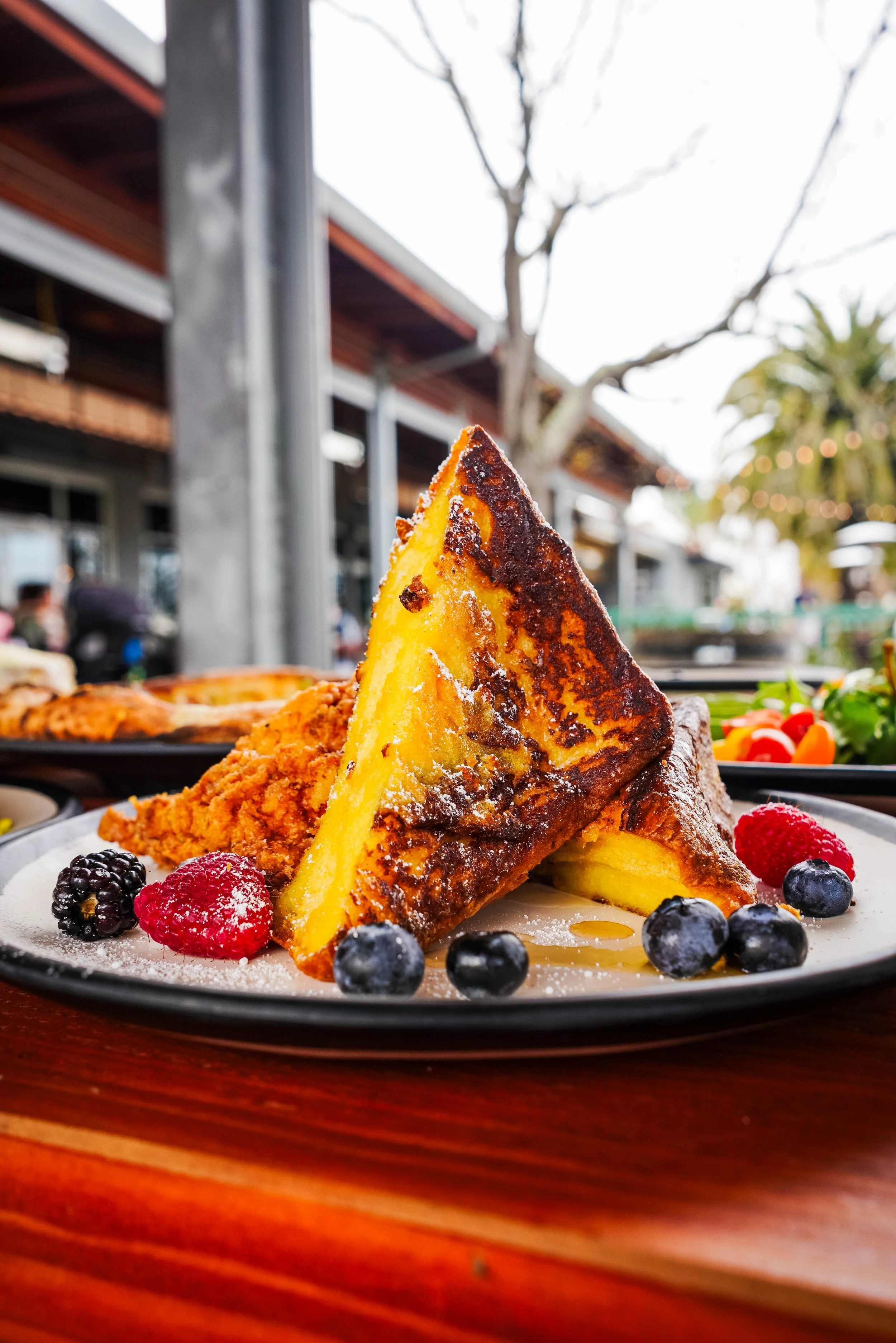 Close-up of French toast topped with powdered sugar, served with fresh berries like blueberries and raspberries, on a round white plate. In the background, outdoor restaurant or cafe setting with blurred trees and building.