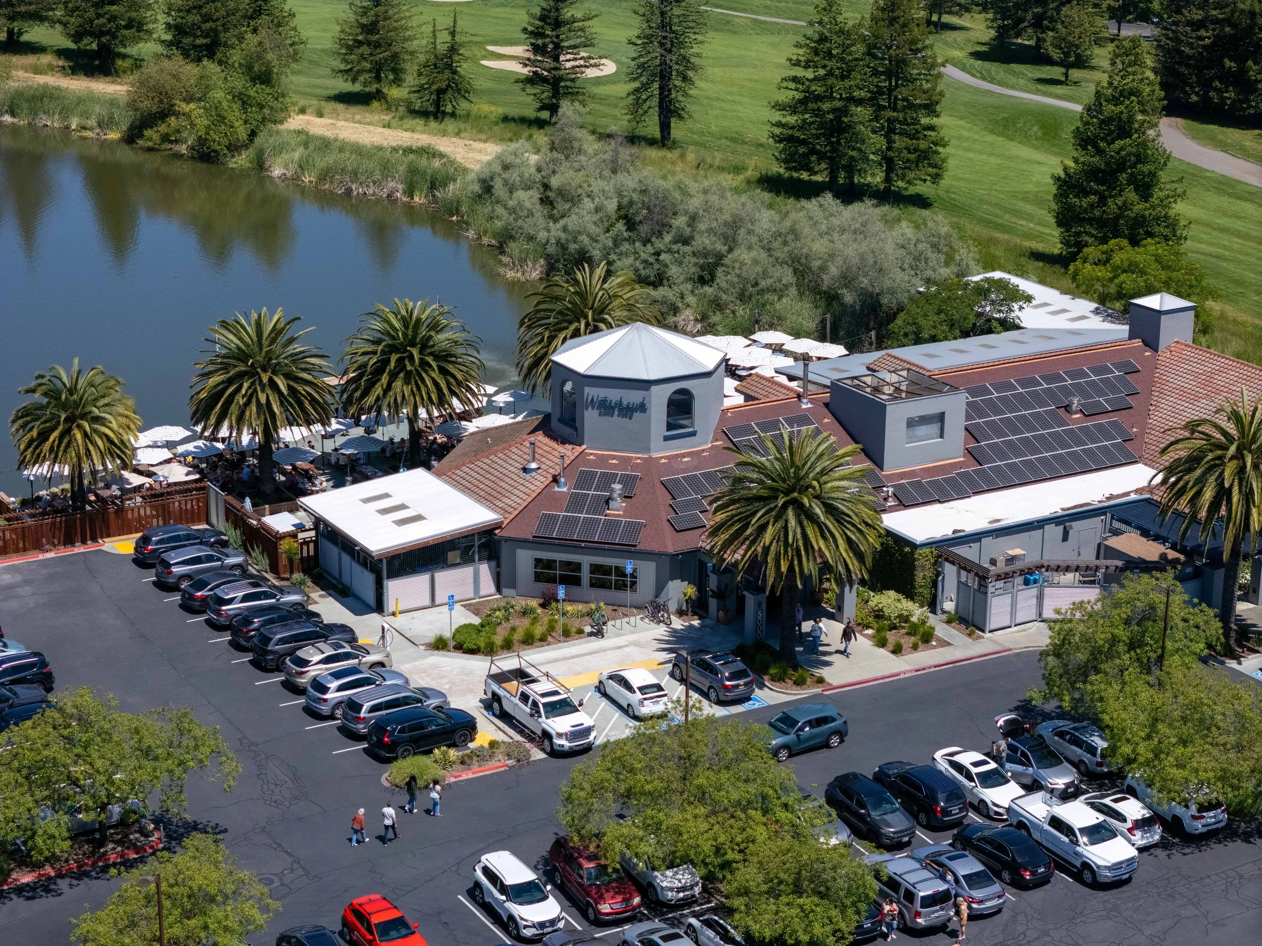 Aerial view of a restaurant with outdoor seating beside a body of water, surrounded by green trees and a parking lot with numerous cars.