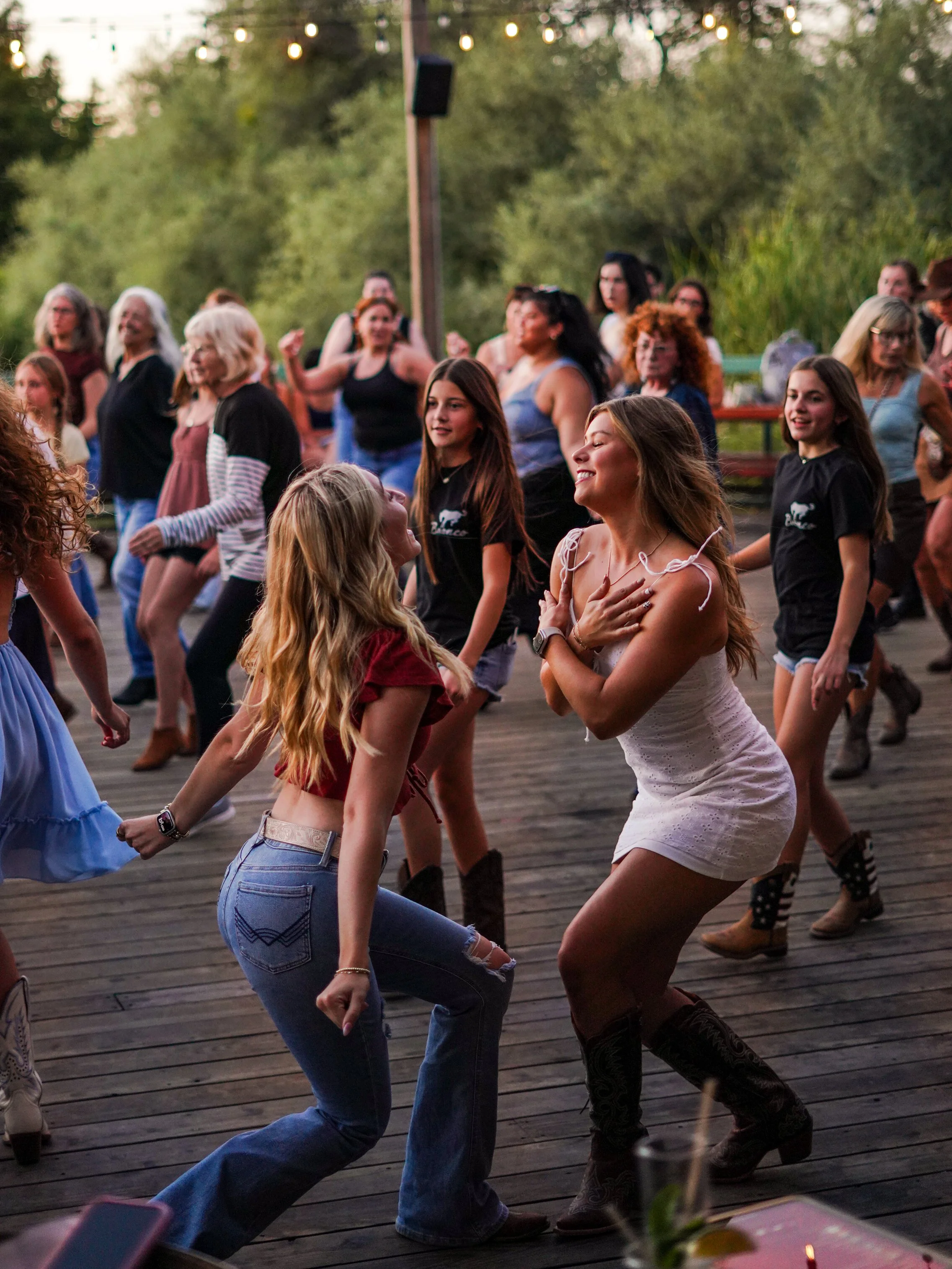 People dancing outdoors on a wooden deck during the evening, with string lights overhead and a lush green background.