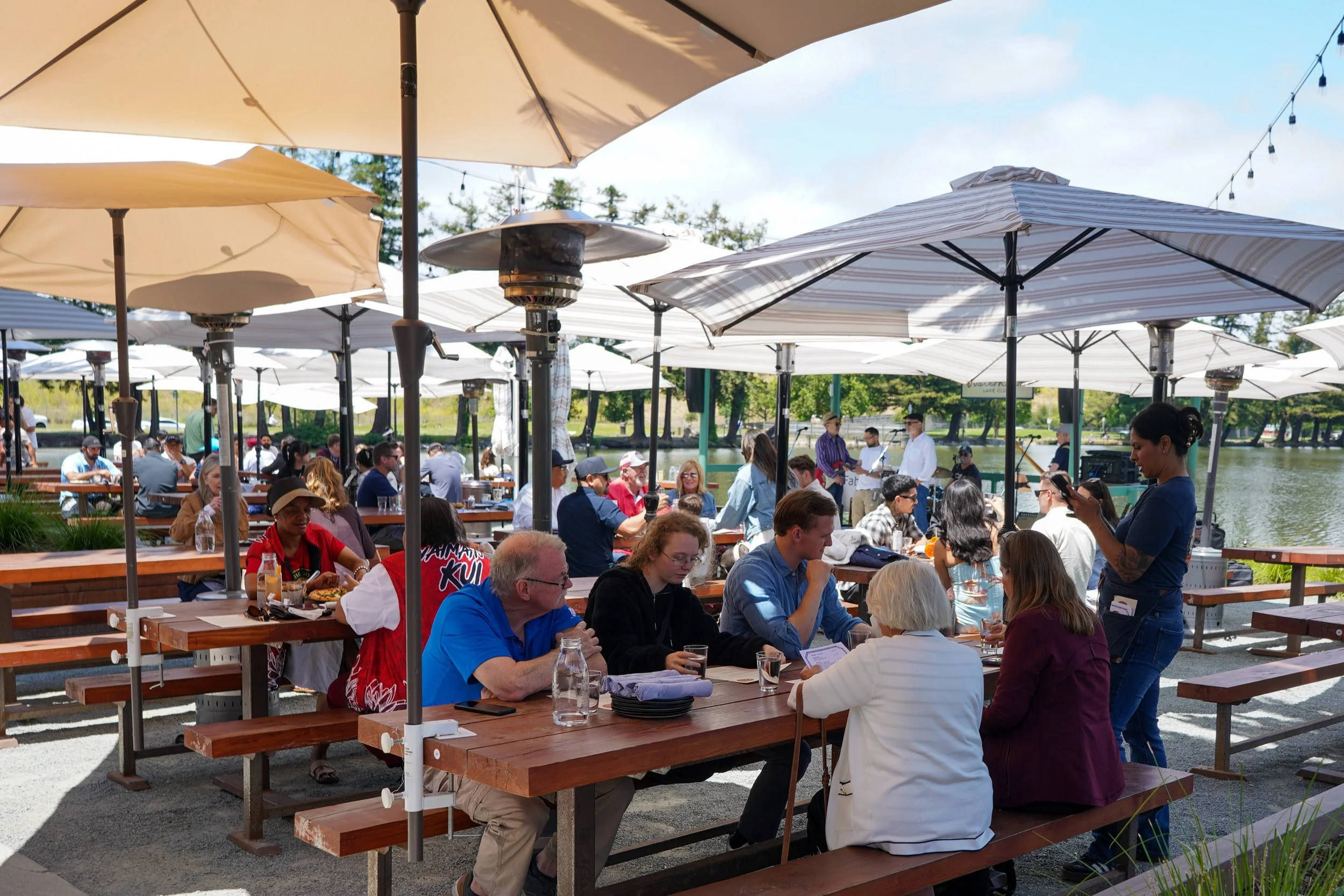 People dining outdoors under umbrellas at a waterfront restaurant on a sunny day.