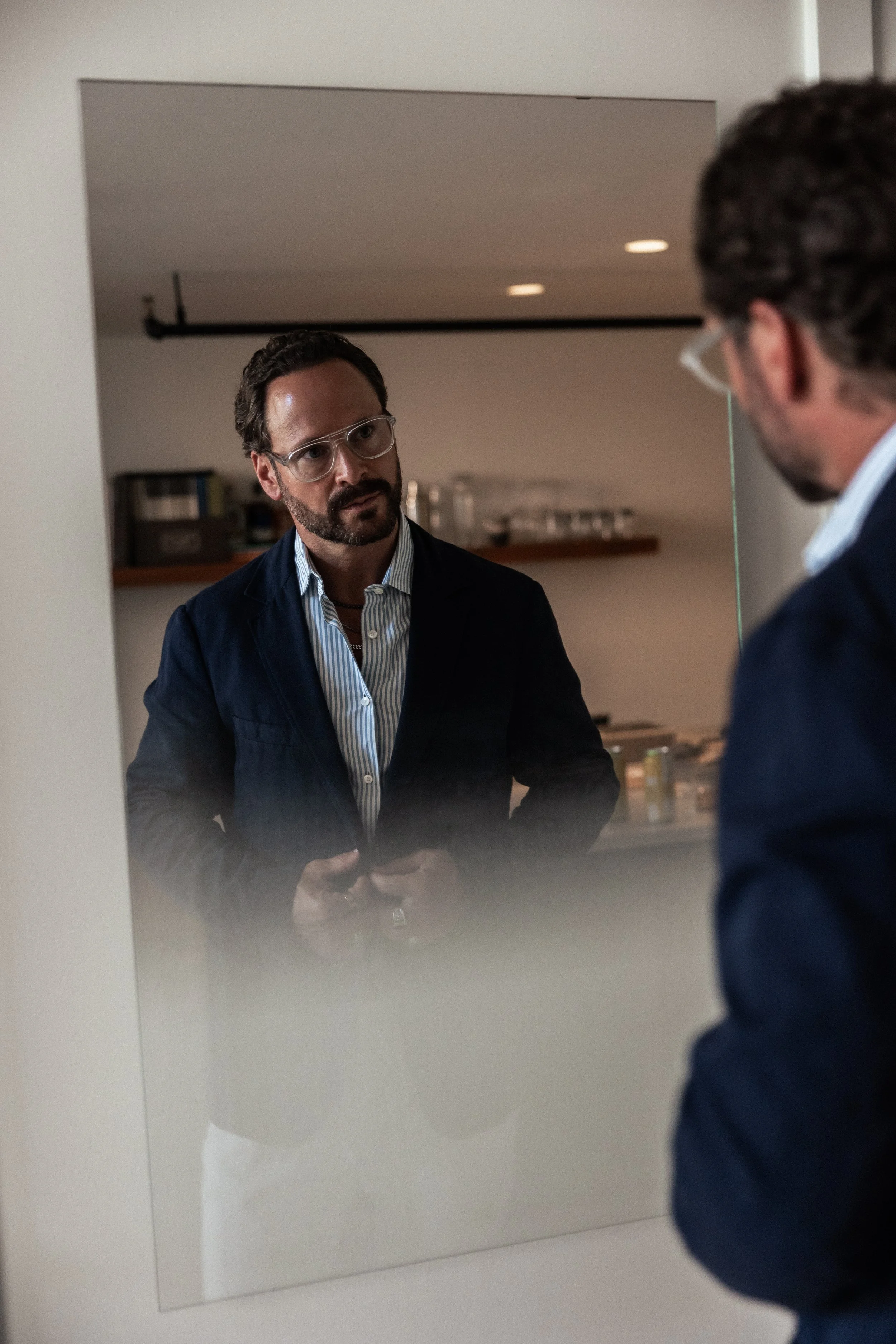 A man with dark hair, beard, glasses, wearing a patterned shirt and dark blazer, is looking at his reflection in a mirror while holding a phone. The reflection shows his face and upper body. Shaun Austin Ford, interior designer.