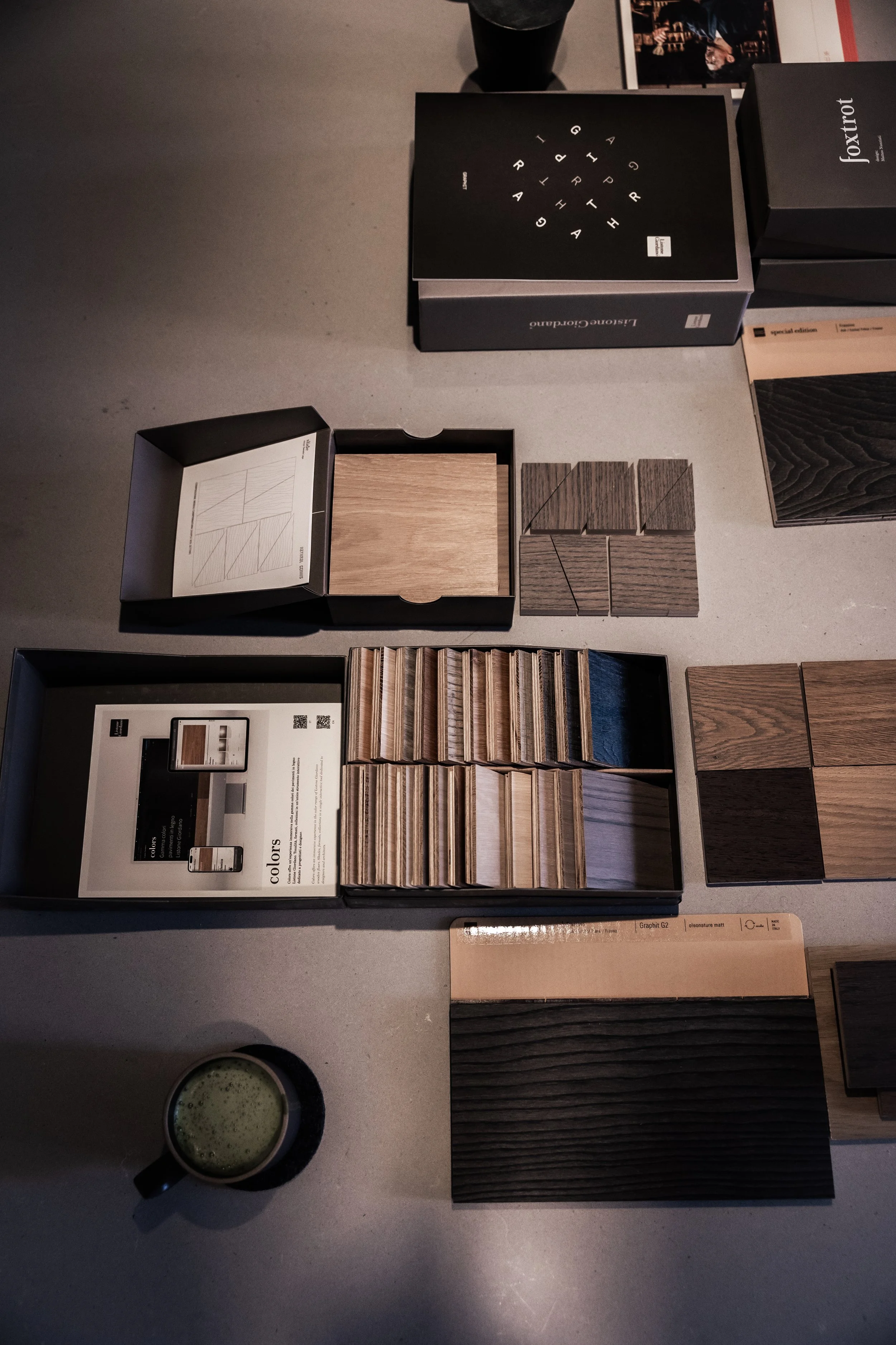 Sample display of various wood and laminate flooring samples laid out on a table, with some boxes and a cup of greenish beverage nearby. Interior design firm, Shaun Ford and Company's material selection for a home renovation project.