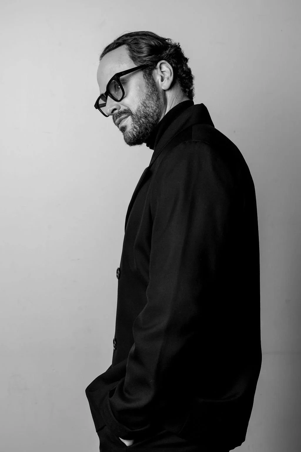 Black-and-white portrait of a man with glasses, a beard, and wavy hair styled back, wearing a dark coat with hands in pockets, against a plain background. Shaun Ford of Shaun Ford & Company portrait for the Shaun Austin Ford digital content creator