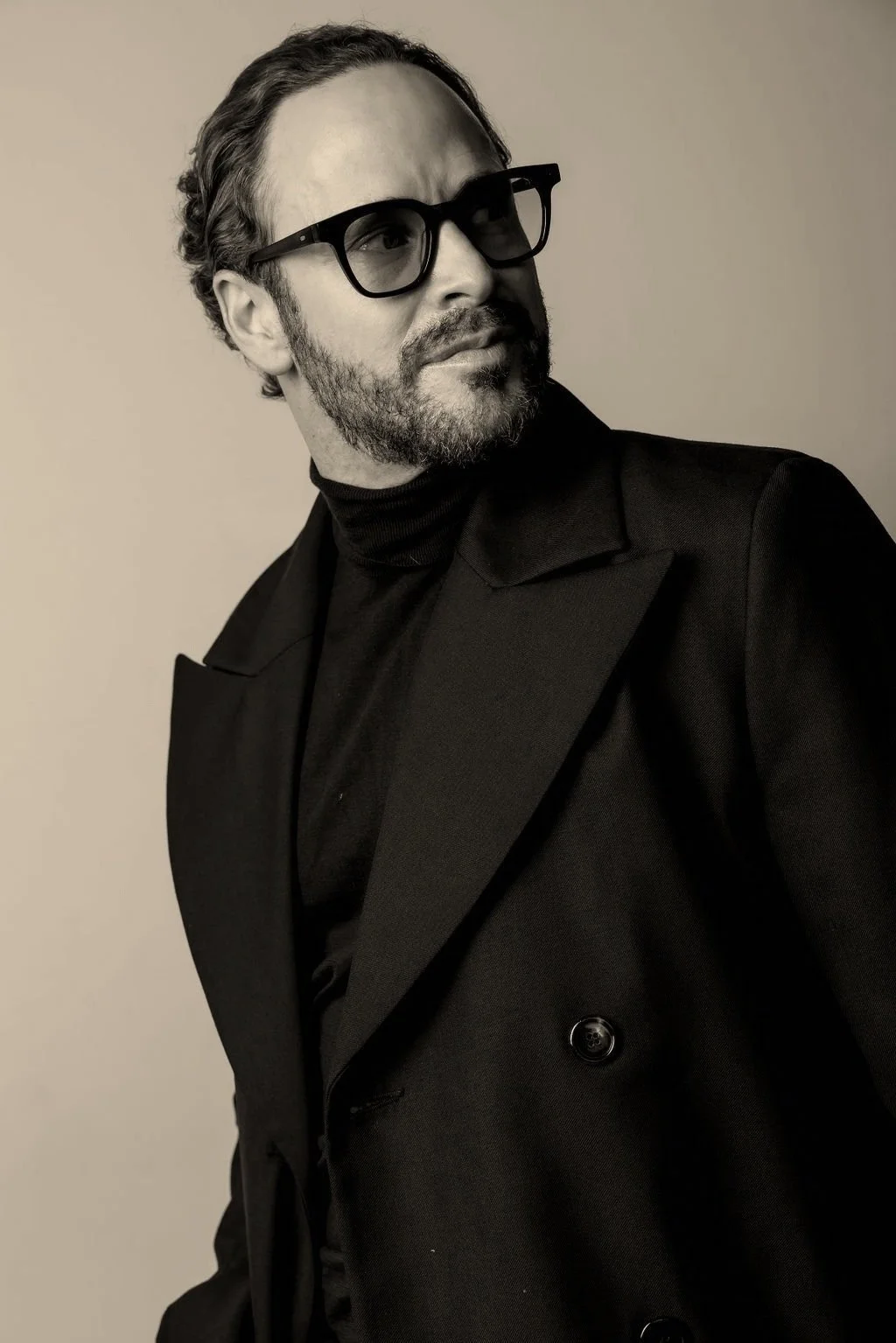 Black and white portrait of a man wearing black glasses, a black turtleneck, and a black blazer with wide lapels, looking to his right with a thoughtful expression.