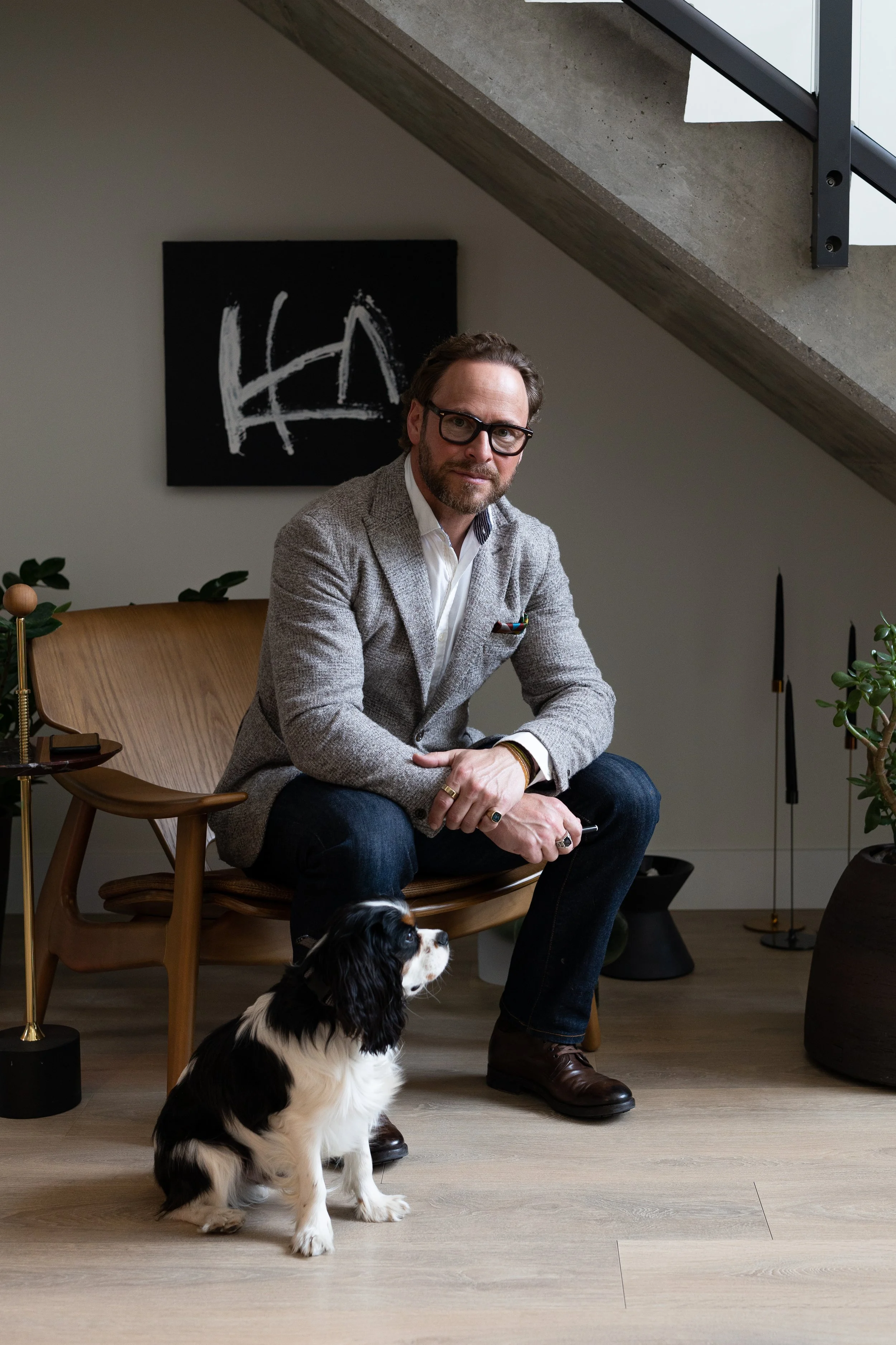 Shaun Ford, wearing glasses and a blazer, sits on a wooden chair in a modern living room with a small dog sitting on the floor nearby. A black-and-white abstract painting hangs on the wall, and indoor plants sit on either side of him.