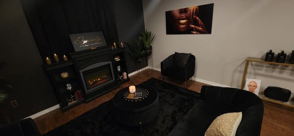 Cozy modern lounge with black furniture, a fireplace, a large wall art of a close-up of a woman's face, and shelves holding candles and skincare products.