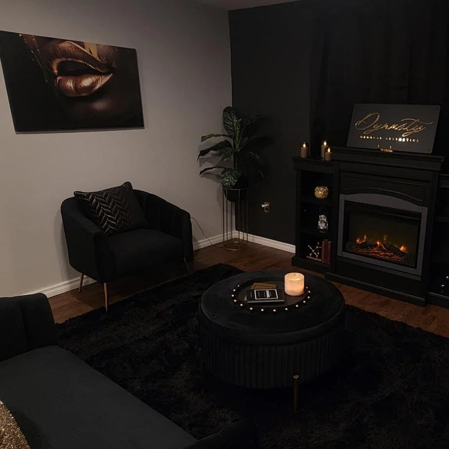 A modern living room with black furniture, a black fireplace, dark wall, tall green plant, and black curtains. Decorations include candles and a framed sign on the fireplace, with elegant gold accents. The room features a candle on the round black ottoman and a black area rug.