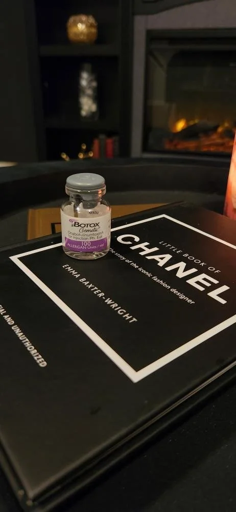 A small vial of Botox cosmetic on top of a Chanel book about the history of the iconic fashion designer, placed on a black surface with a fireplace and shelves in the background.