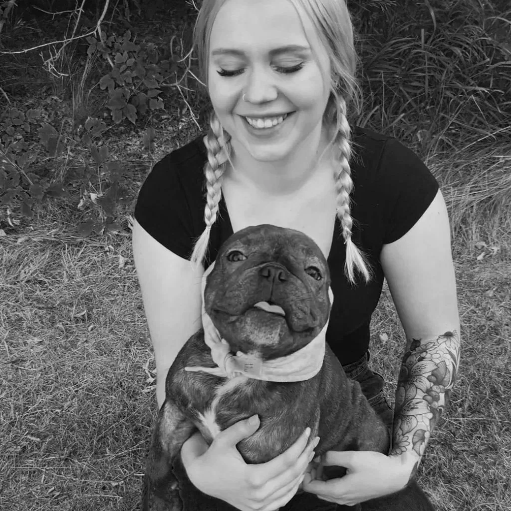 A smiling woman with braided hair and tattoos on her arm holding a large dog outdoors.