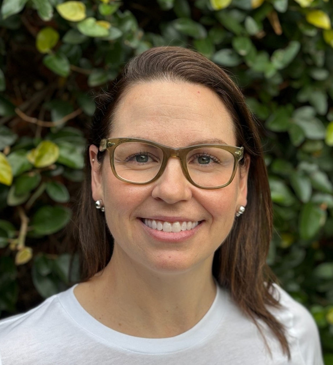 A smiling woman with brown hair, glasses, and pearl earrings standing outdoors with green foliage in the background.