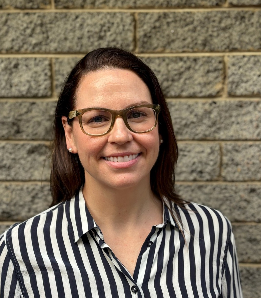 A woman with brown hair, wearing glasses, a striped black and white blouse, and pearl earrings, smiling in front of a brick wall.