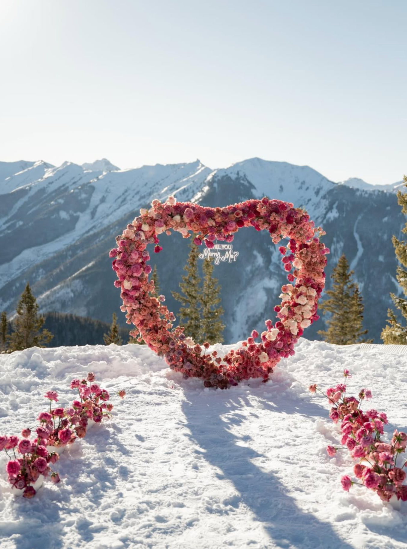 A heart-shaped arch made of pink and white flowers in a snowy landscape with mountains in the background, and a sign hanging inside the heart reading 'Will You Marry Me?'
