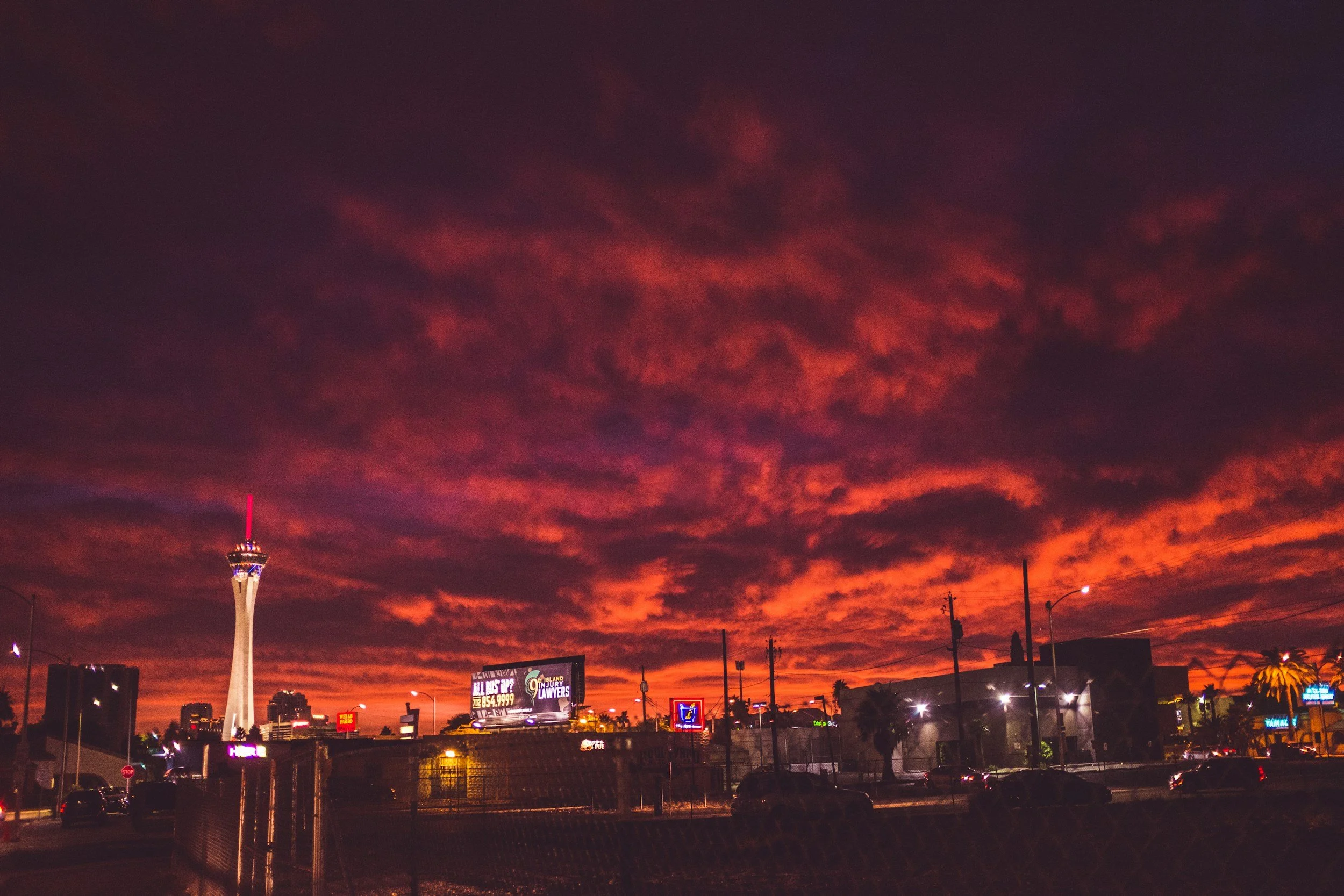 Las Vegas skyline at sunset with dark red and purple clouds, visible satellite tower, billboards, streetlights, and palm trees.