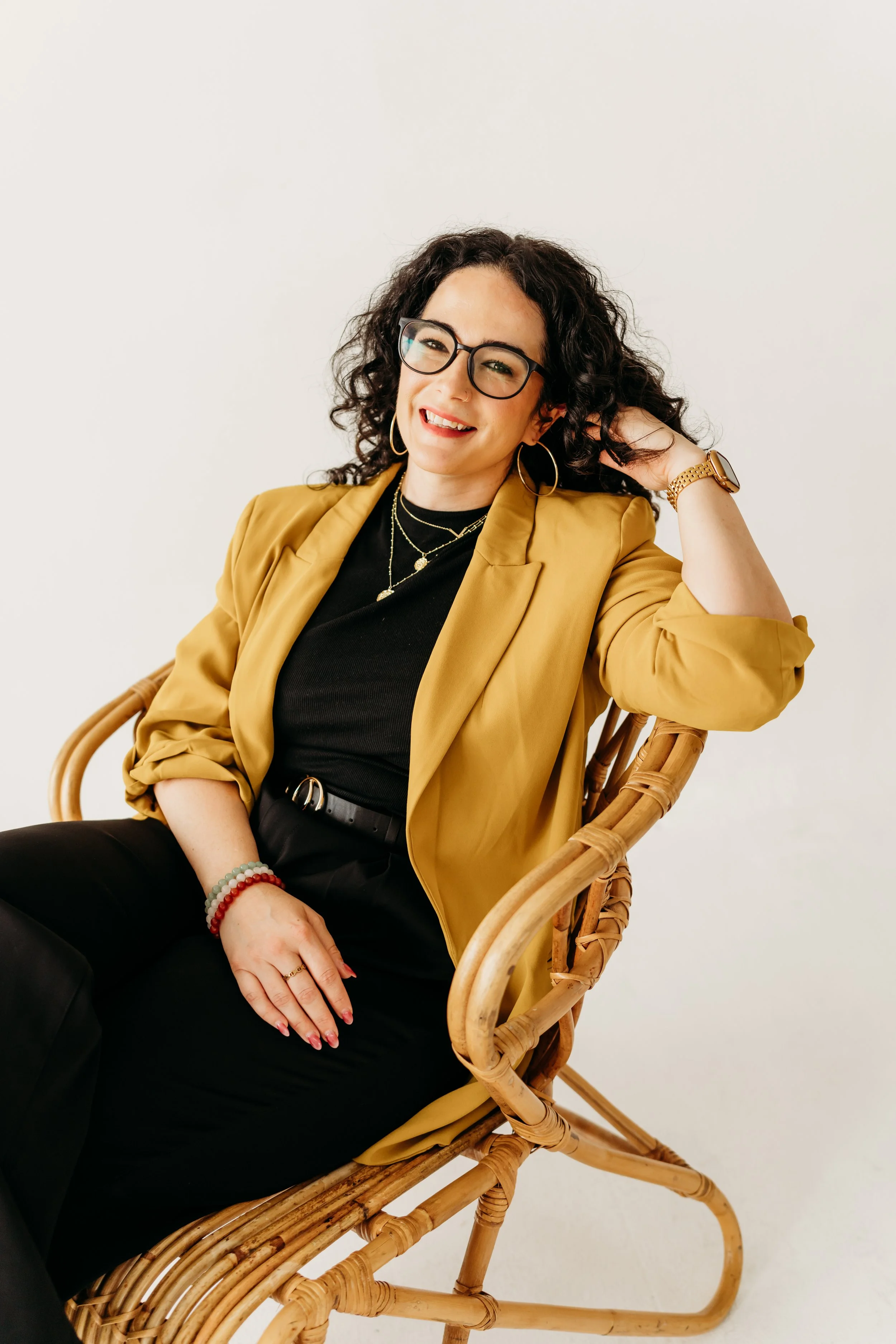 Carly Shadid, LMFT, smiling in a mustard blazer in a rattan chair