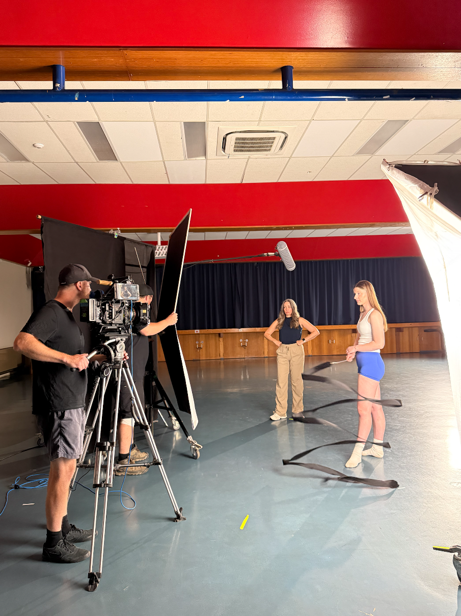Film crew setting up for a shoot with two women in a studio, one in a white top and blue shorts, and another in a black top and beige pants.