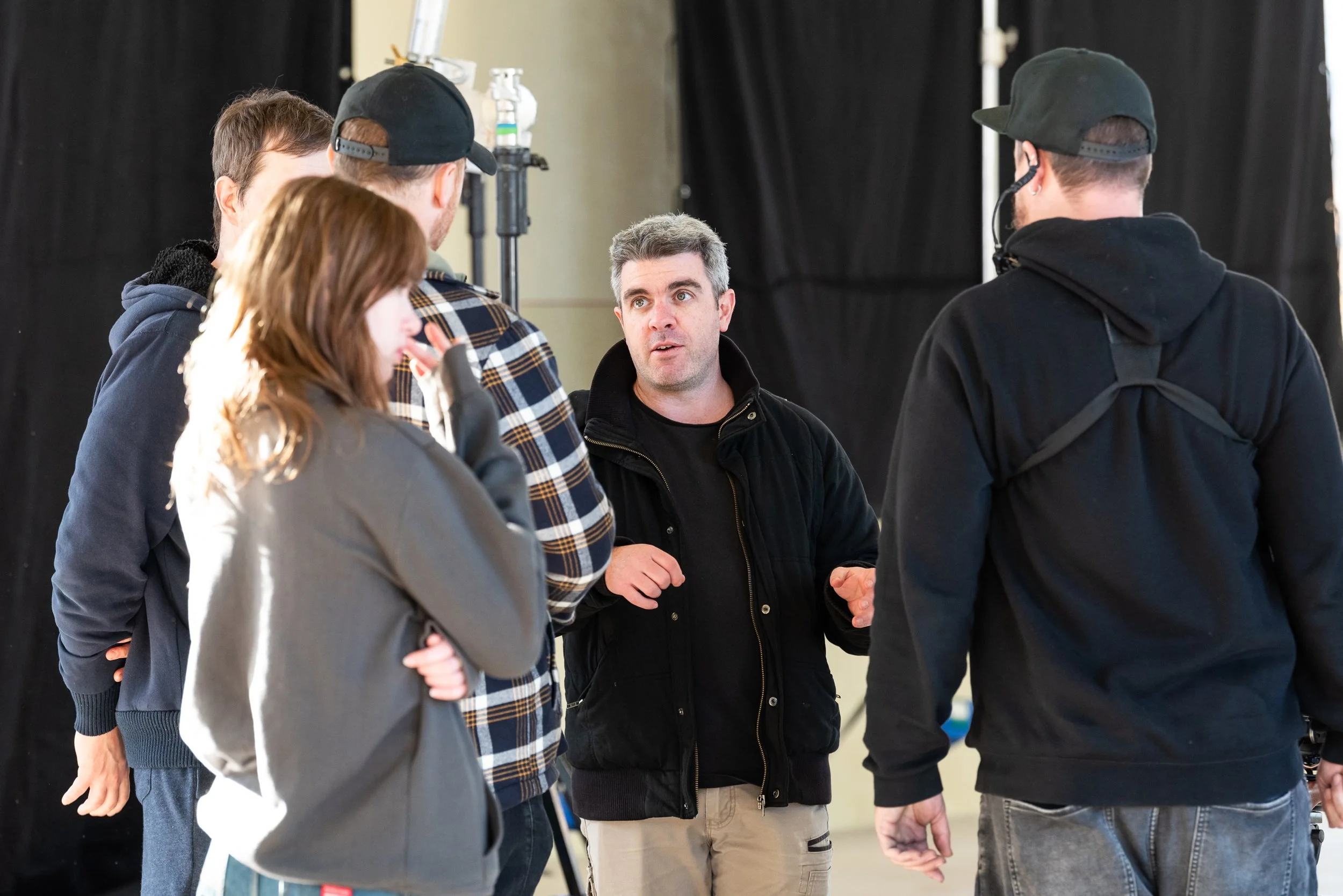 A group of five people are gathered in a conversation, with one man speaking and gesturing with his hands, while the others listen. They are in an indoor setting with black curtains or backdrop.