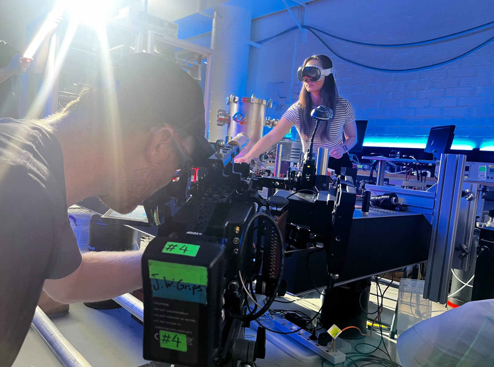A woman wearing virtual reality goggles and headphones working at a science or technology laboratory table with scientific equipment, with a man operating a camera or recording device in the foreground.