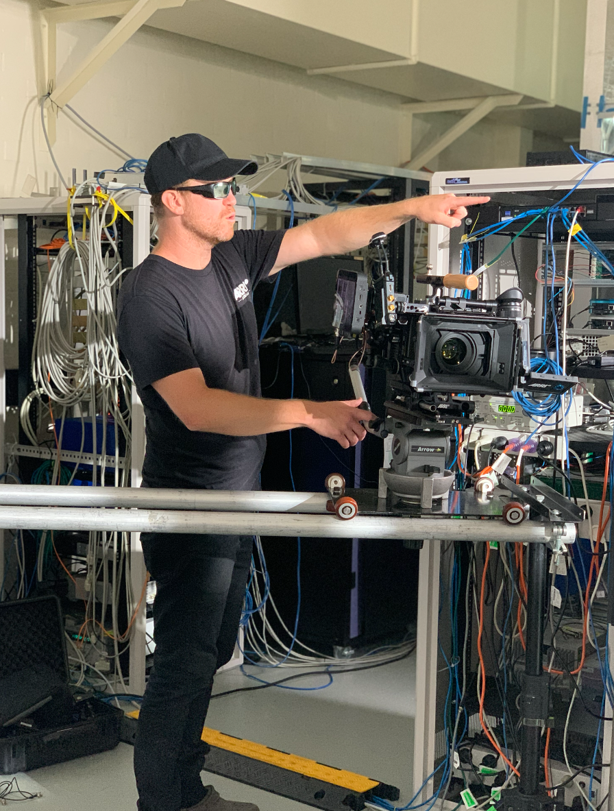 A man wearing sunglasses, a black cap, and a black T-shirt operating a professional video camera on a stabilizer rig in a room with numerous cables and electronic equipment.