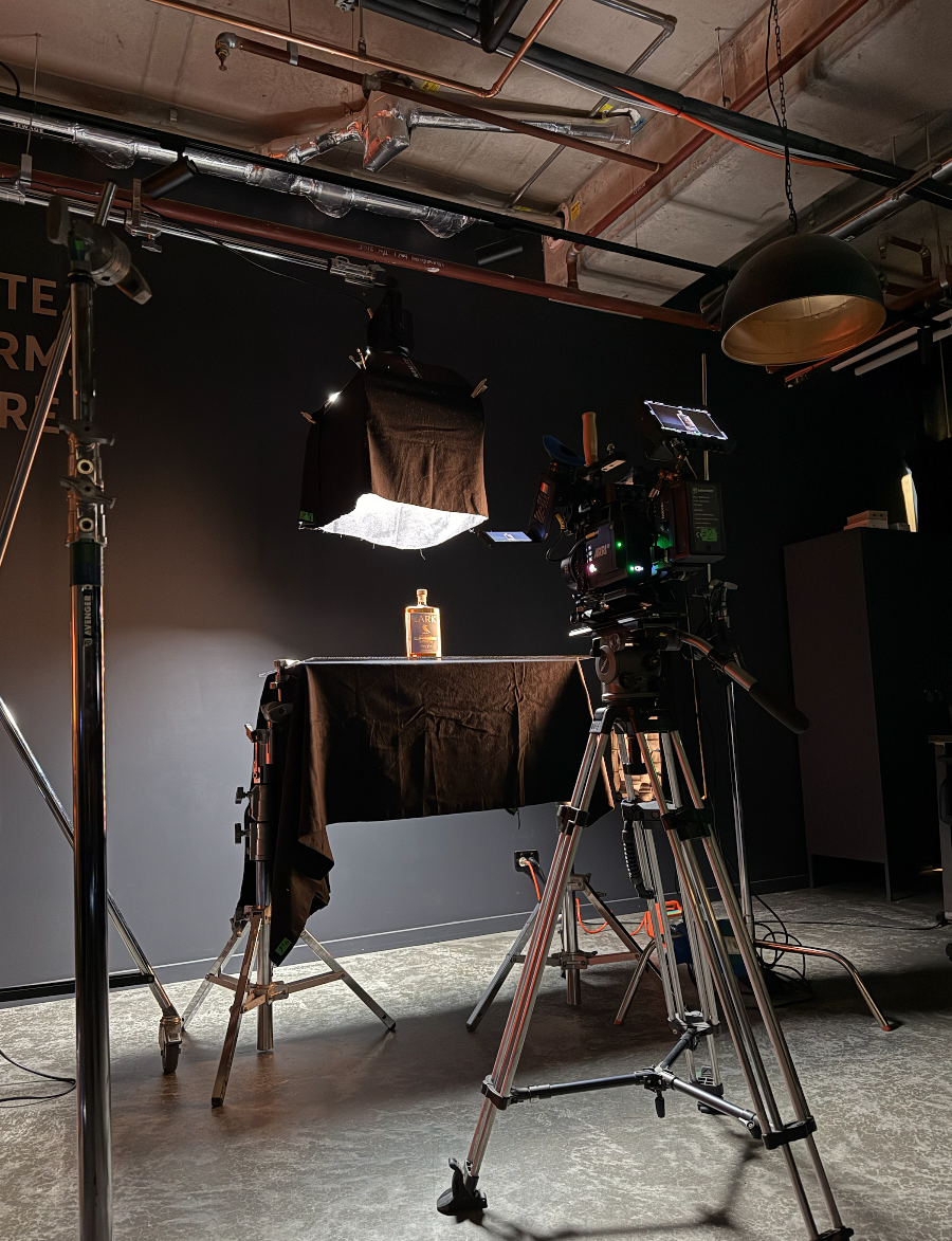 A professional filming setup for a product shot, with a camera aimed at a liquor bottle on a black table, illuminated by studio lights, in a dark room with black walls and equipment.