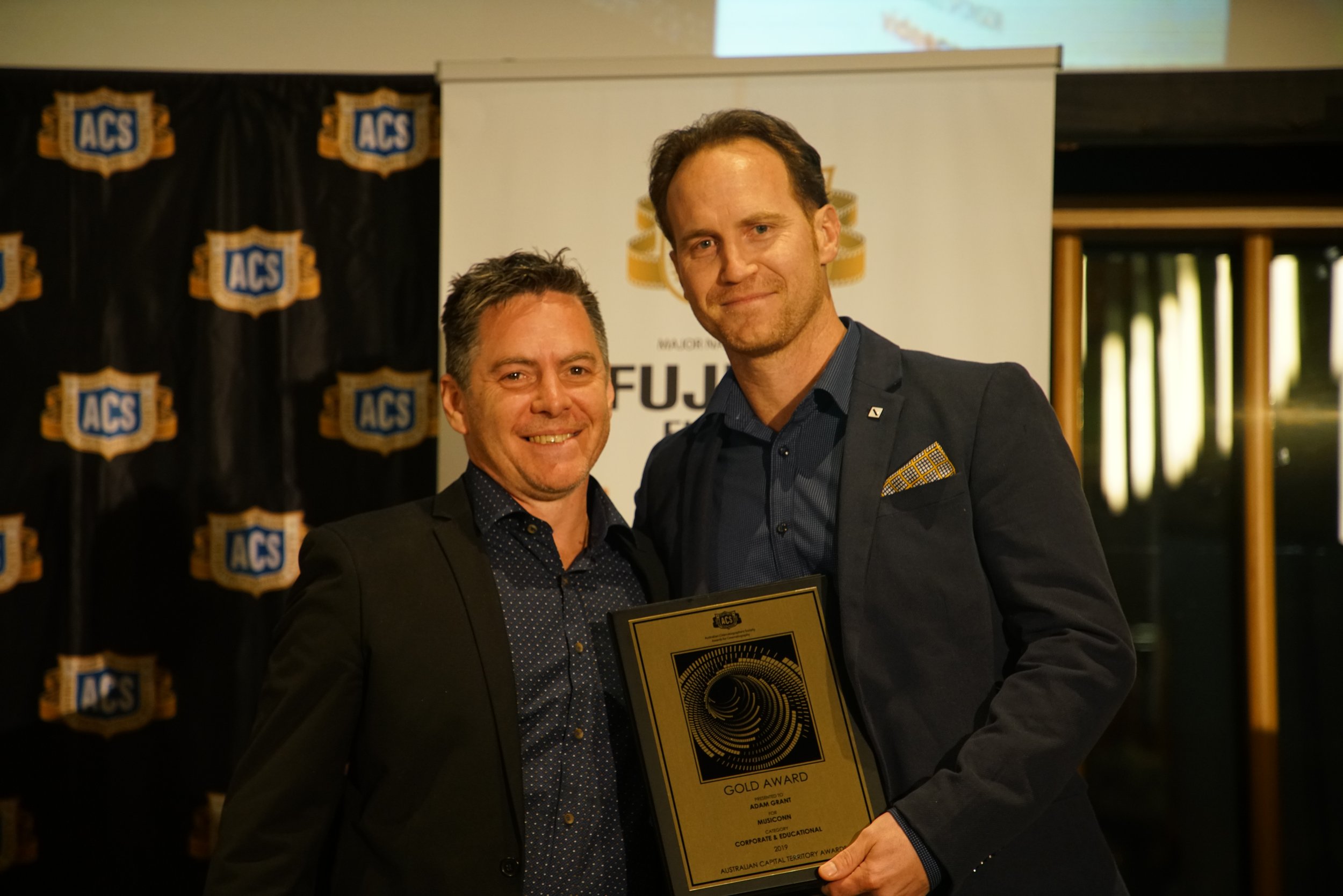 Two men dressed in formal attire, smiling at the camera, at an awards event. The man on the right holds a gold award plaque with text, and the background features a banner with the ACS logo.