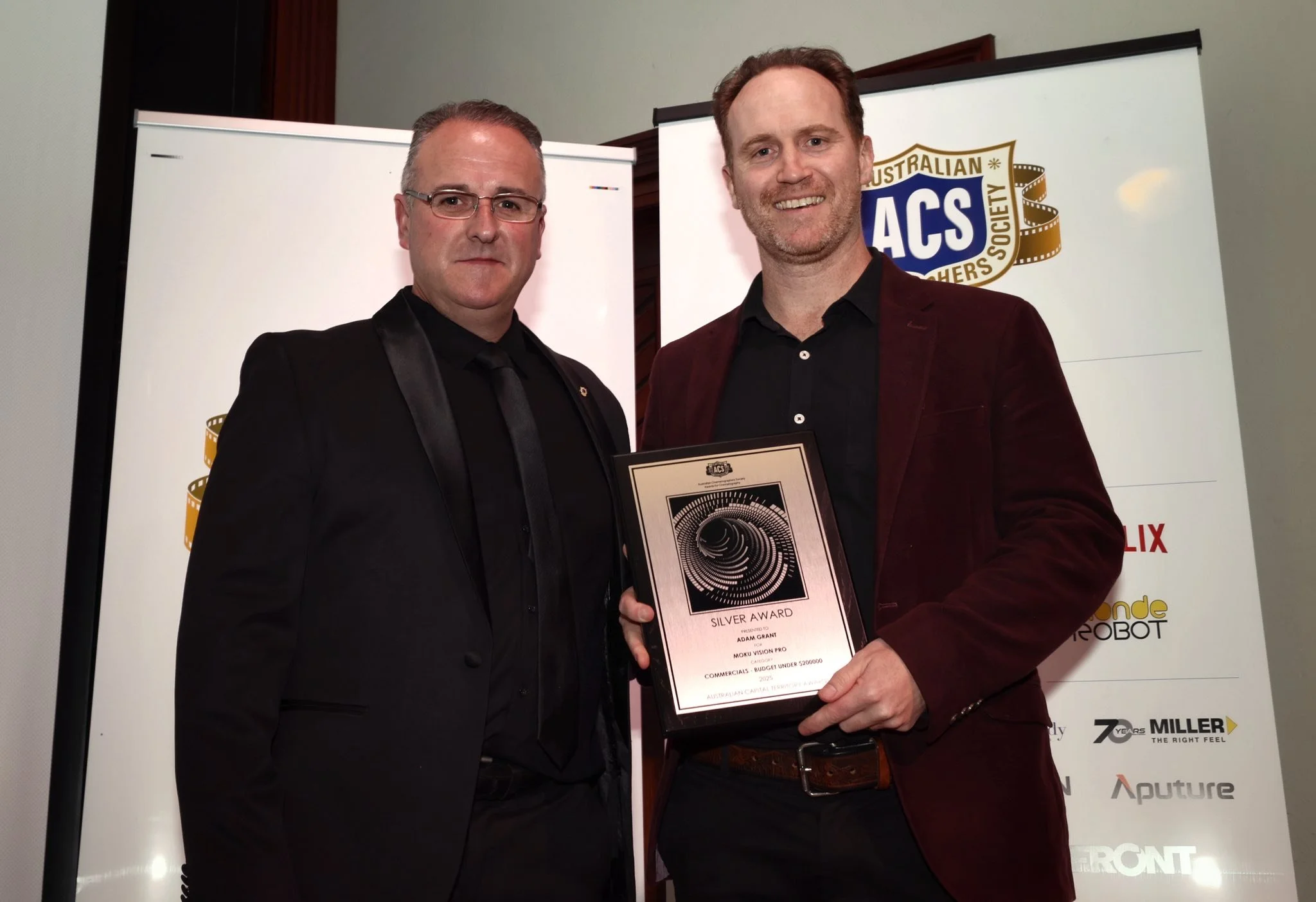 Two men in formal attire standing together at an awards event, holding a certificate and smiling, with a backdrop featuring logos including the Australian Cinematographers Society and Netflix.