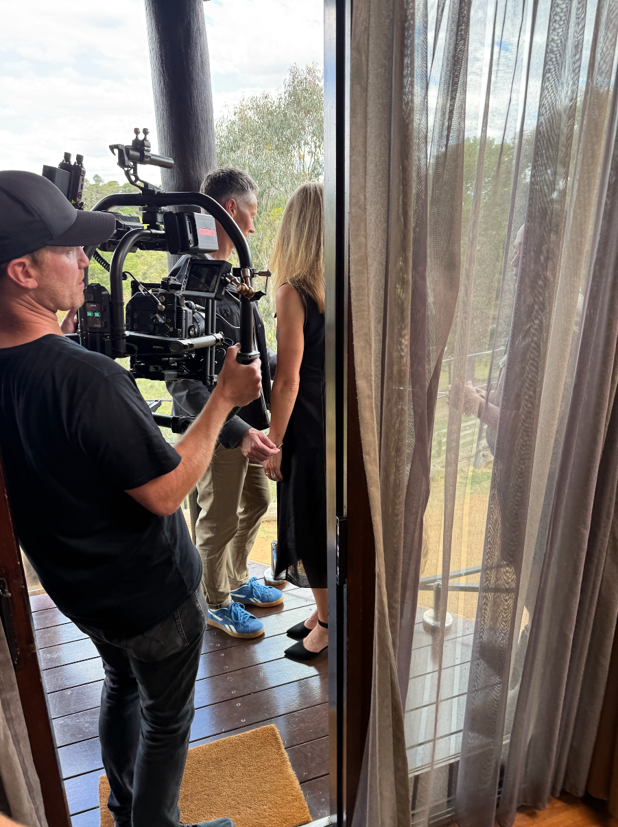 A film crew and director are filming a scene with a woman in a black dress on a balcony, with trees and a cloudy sky in the background.