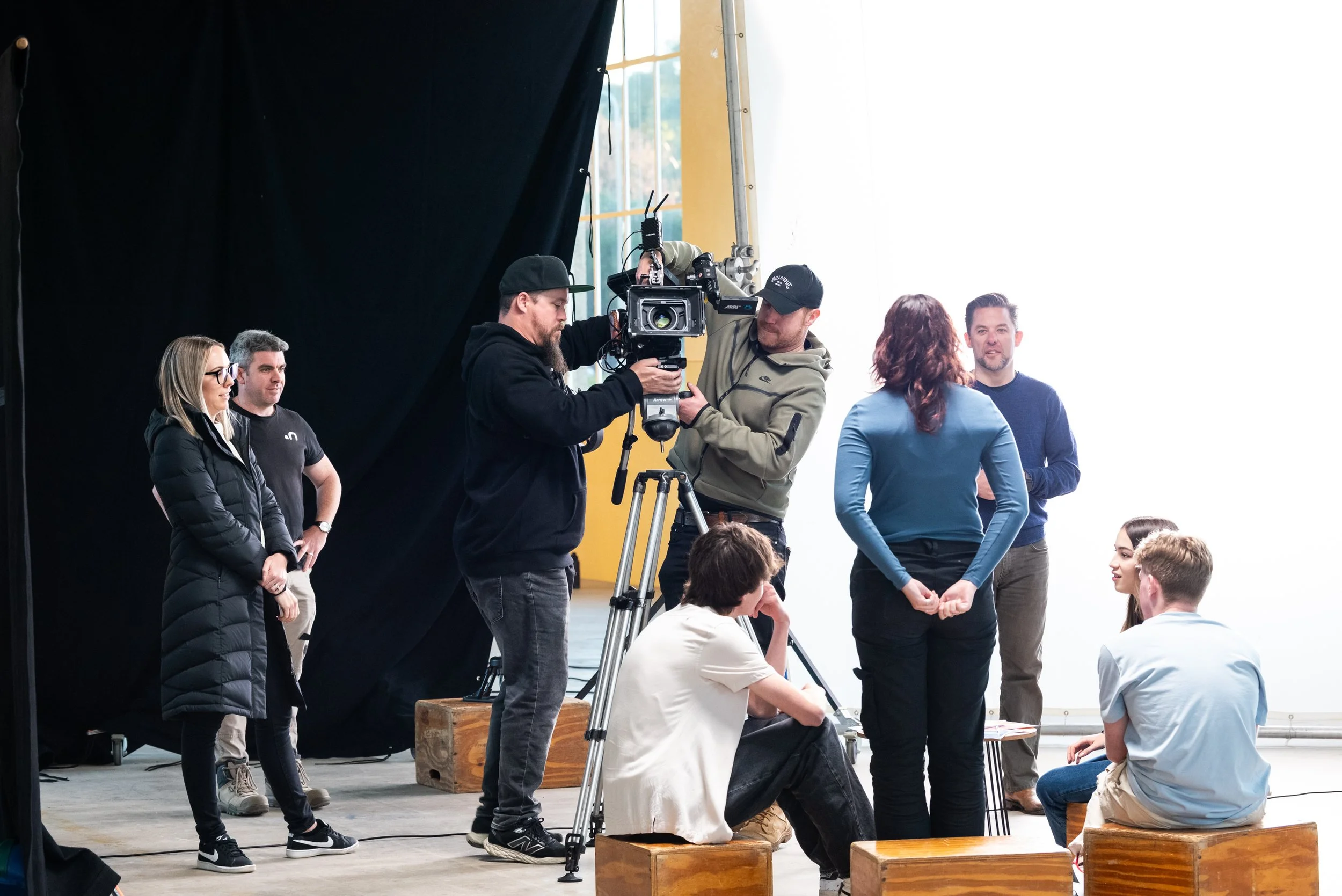 Film crew filming a scene with actors in a studio, with camera operators and crew members working around them.