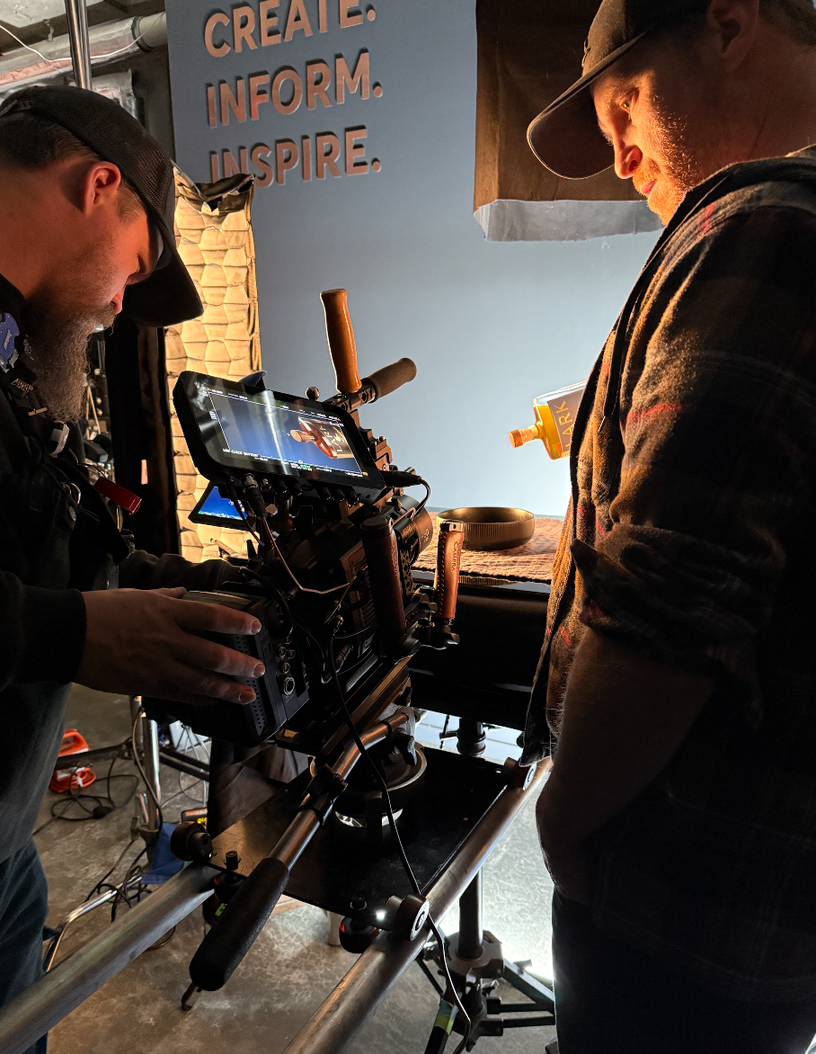 Two men on a film set operating professional camera equipment, with a blue wall behind them displaying the words 'Create. Inform. Inspire.'
