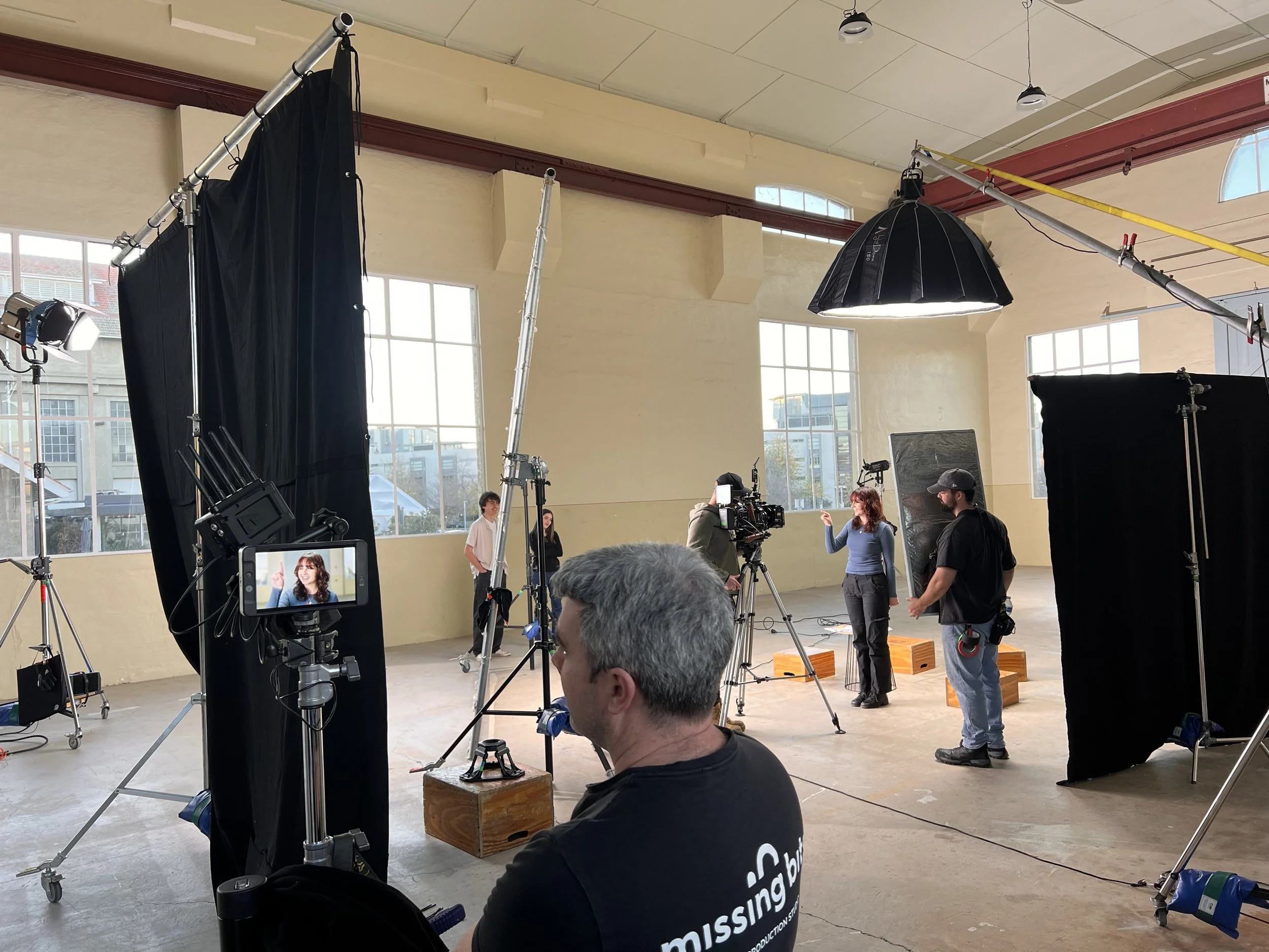 Filming studio with crew setting up shot featuring a woman in a blue sweater talking and gesturing, surrounded by lighting and cameras, with large windows in the background.