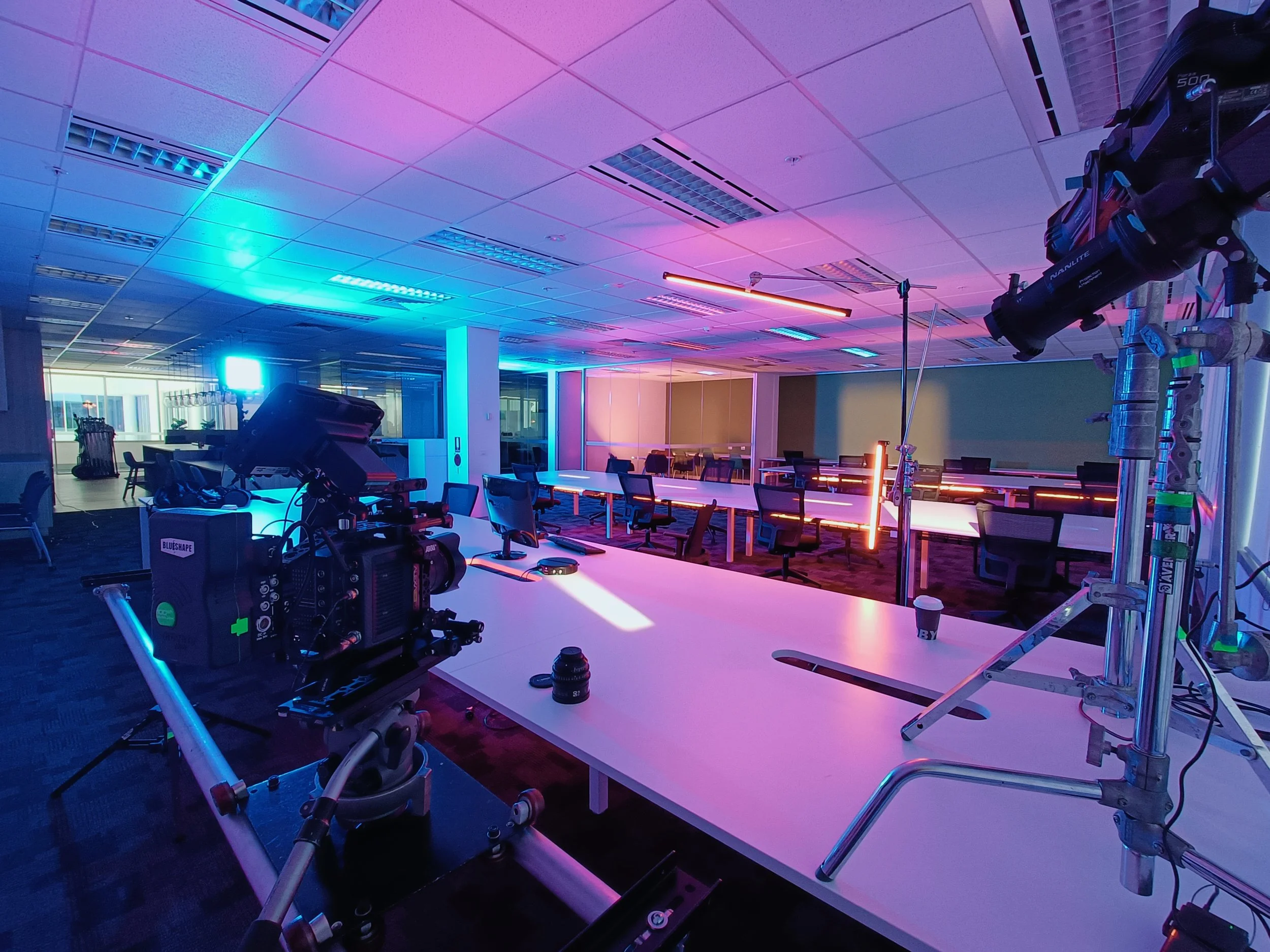 Film or television set in a modern office conference room with cameras and lighting equipment, illuminated by colored blue and pink lights.