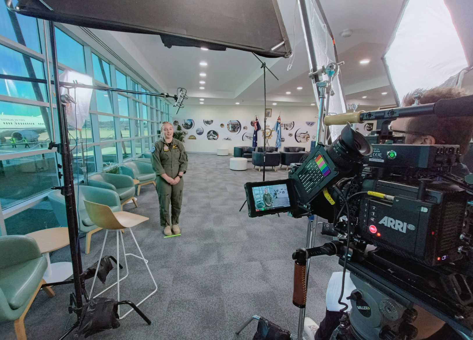 A woman in a military uniform standing in an airport lounge, with aircraft visible through large windows behind her. The image also shows professional filming equipment, including a camera and lights, indicating the scene is part of a video or photo 
